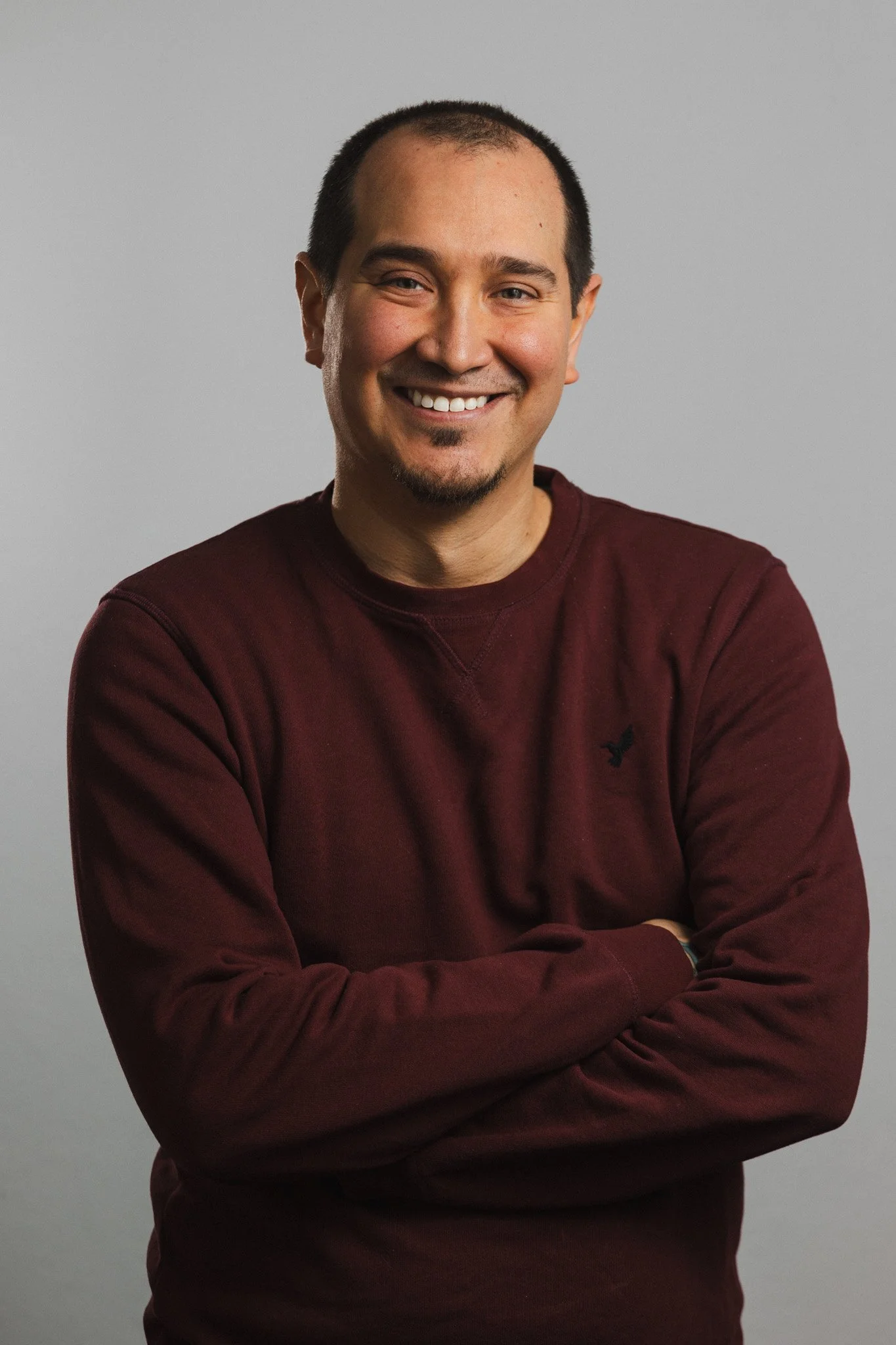 A smiling man with short dark hair, wearing a maroon long-sleeve shirt, standing against a plain gray background.