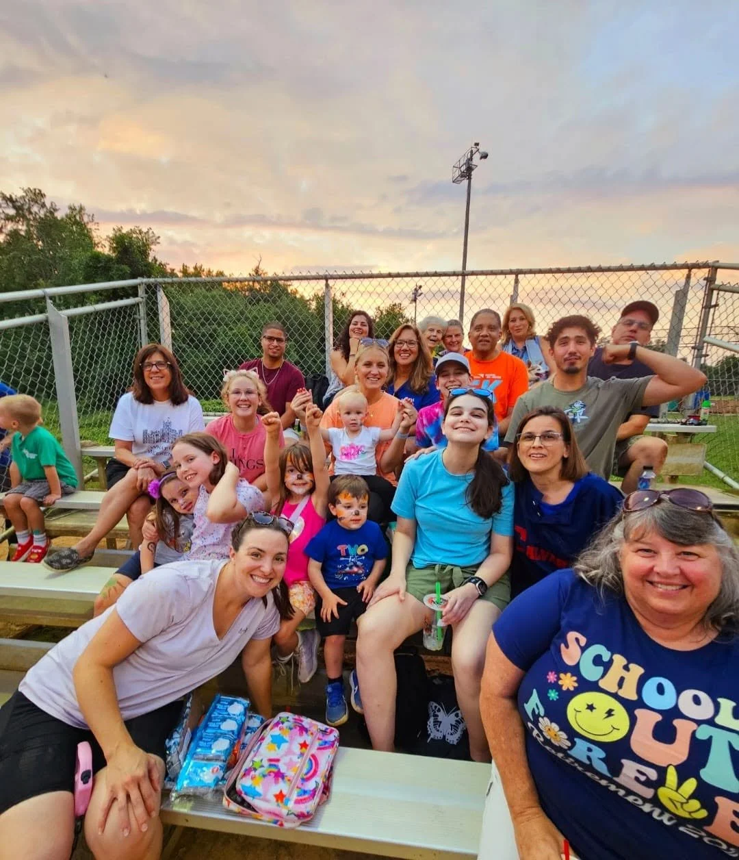 Group of children and adults sitting on bleachers at a baseball field during sunset, smiling and posing for a photo.