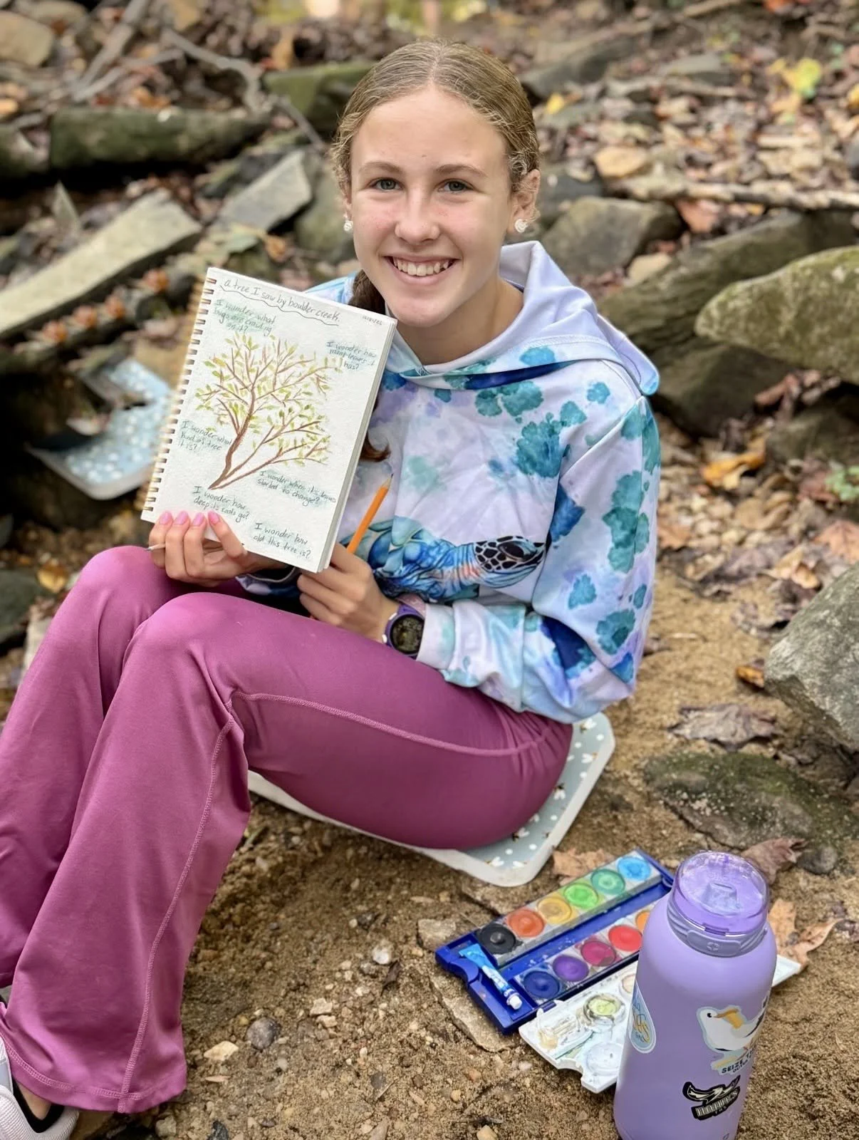 A young girl sitting outdoors on a rock, holding a sketchbook with a drawing of a tree and some notes. She is smiling, wearing a blue and white tie-dye hoodie, pink pants, and a wristwatch. Art supplies, including watercolor paints and a water bottle, are on the ground beside her in a natural setting with fallen leaves and rocks.