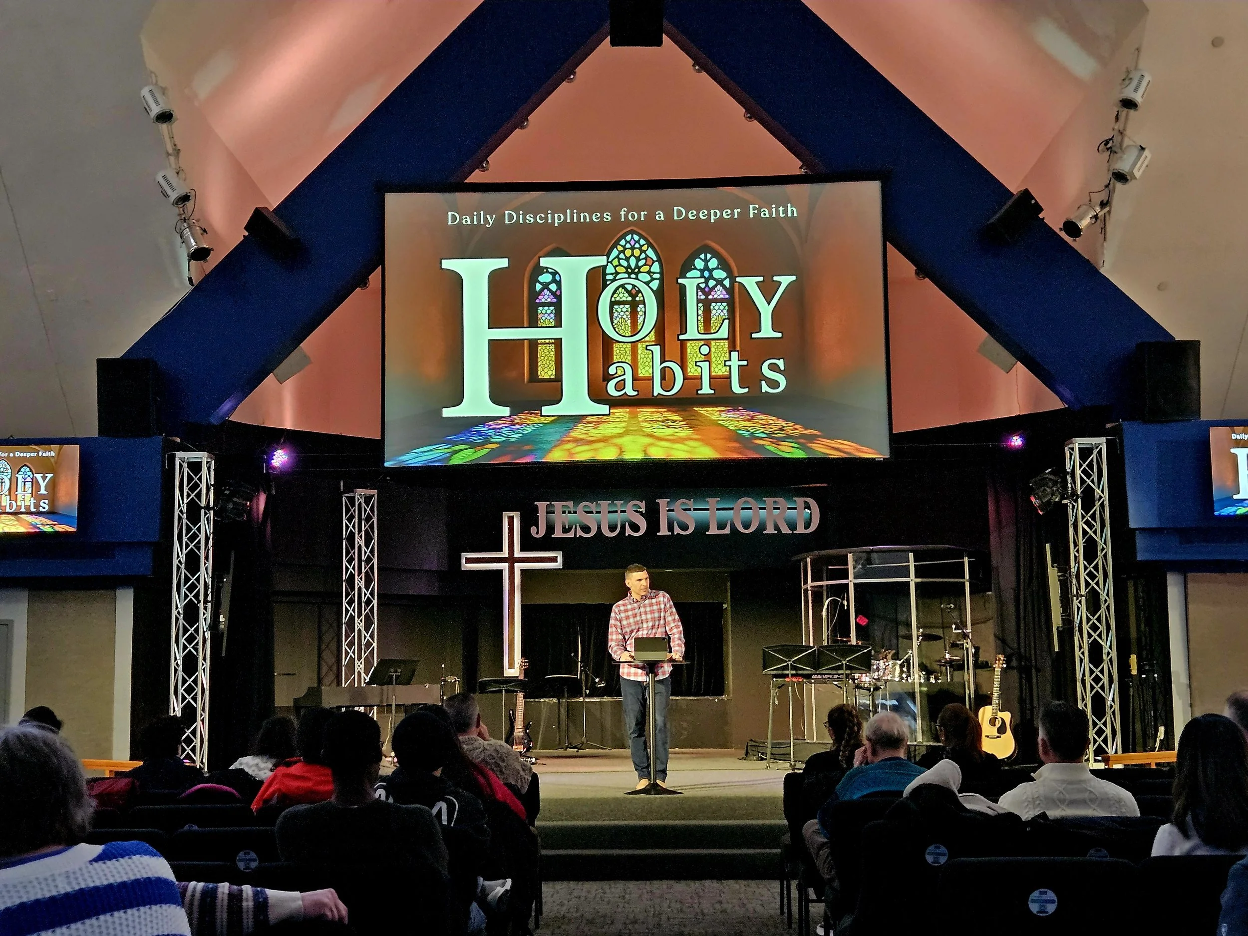A church or worship space with a large screen displaying the words "Daily Disciplines for a Deeper Faith" and "HOLY habits" with stained glass window imagery in the background. A man is speaking at a podium, and there are musical instruments and chairs on the stage. Audience members are seated, some wearing colorful clothing.