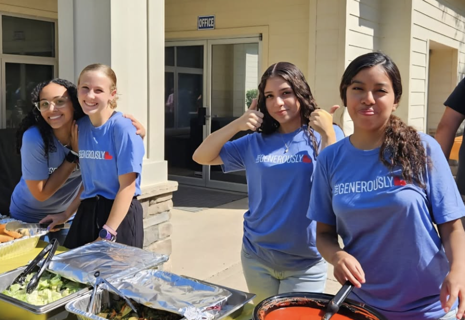 Four young women wearing blue shirts are standing outdoors at a food serving table, smiling and posing for the camera at what appears to be a community event or gathering.