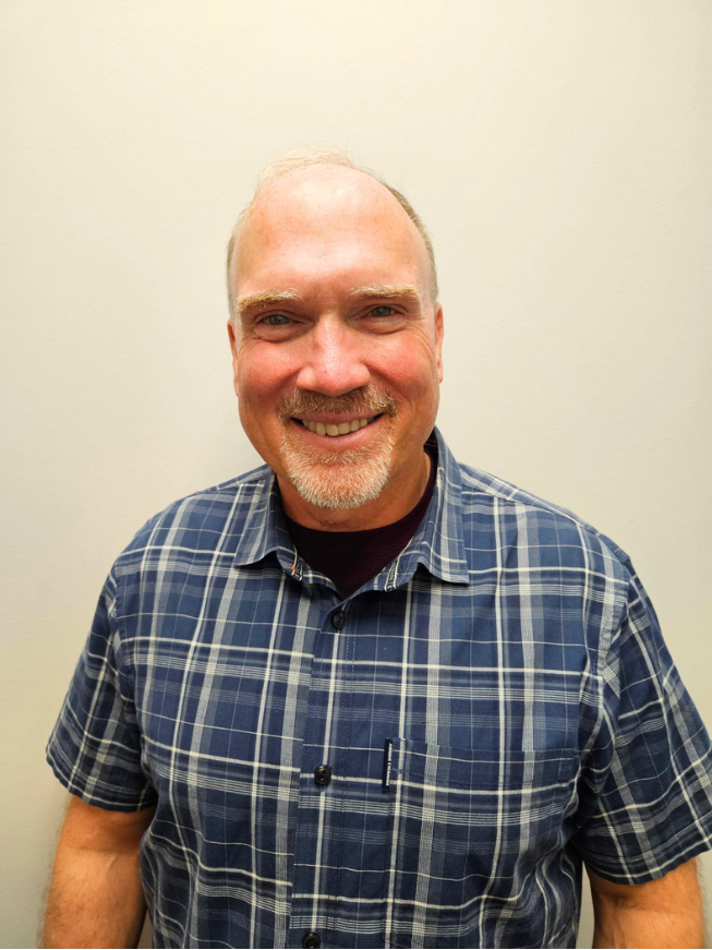 A smiling man with a beard and mustache wearing a blue plaid shirt standing against a plain light-colored wall.