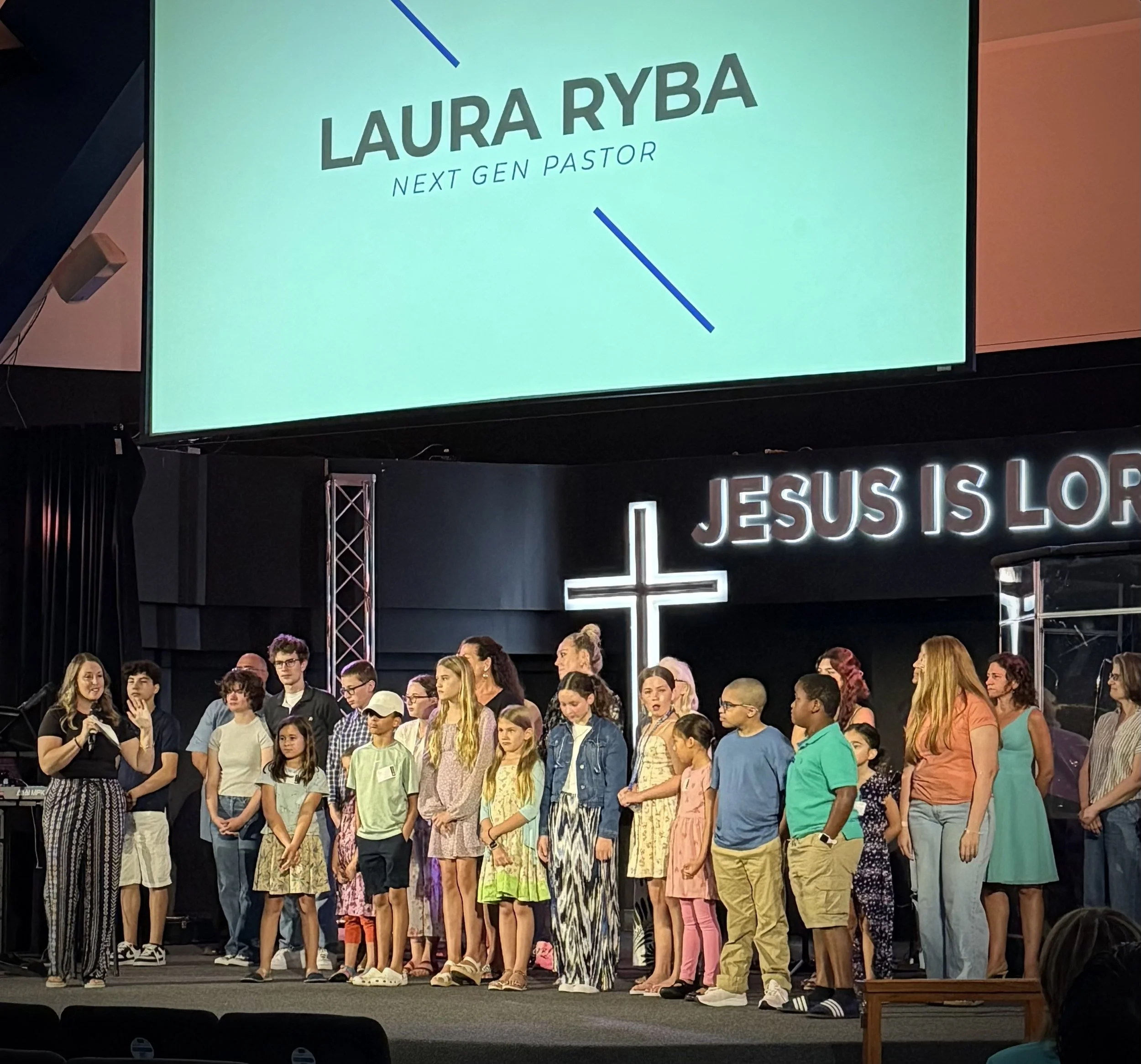 A large group of people, mostly children, standing on stage in front of a sign with the name Laura Ryba and the title Next Gen Pastor. Some are looking at a woman speaking into a microphone, while others are looking down or to the side.