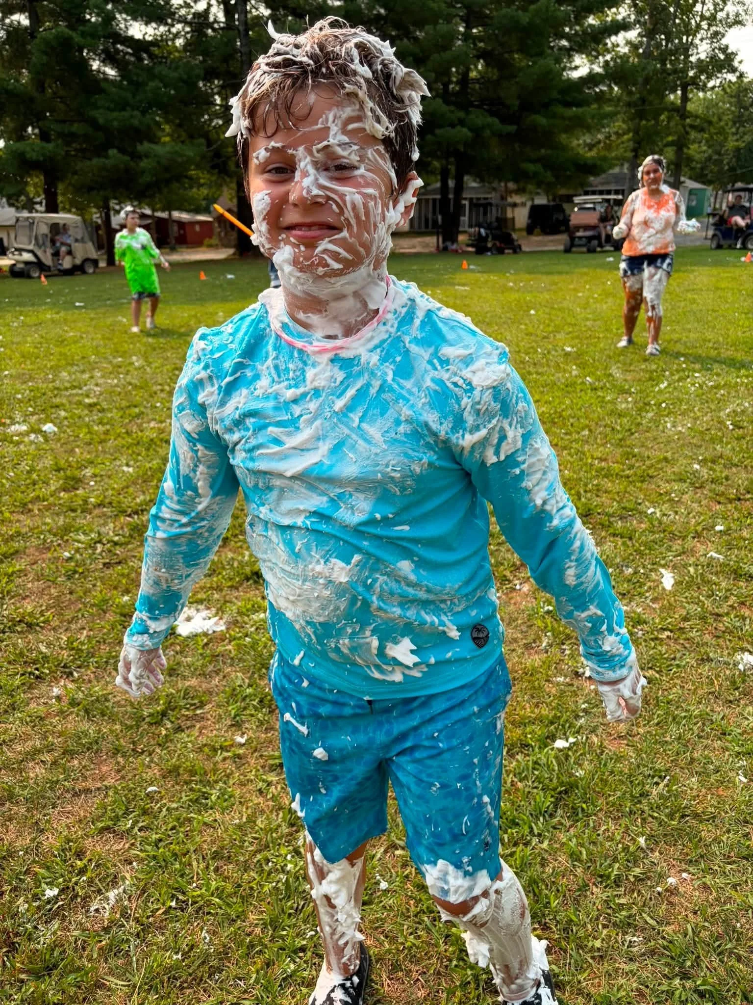 A boy covered in white foam, smiling outdoors on a grassy field during daytime, with other children and adults in the background participating in a playful event.