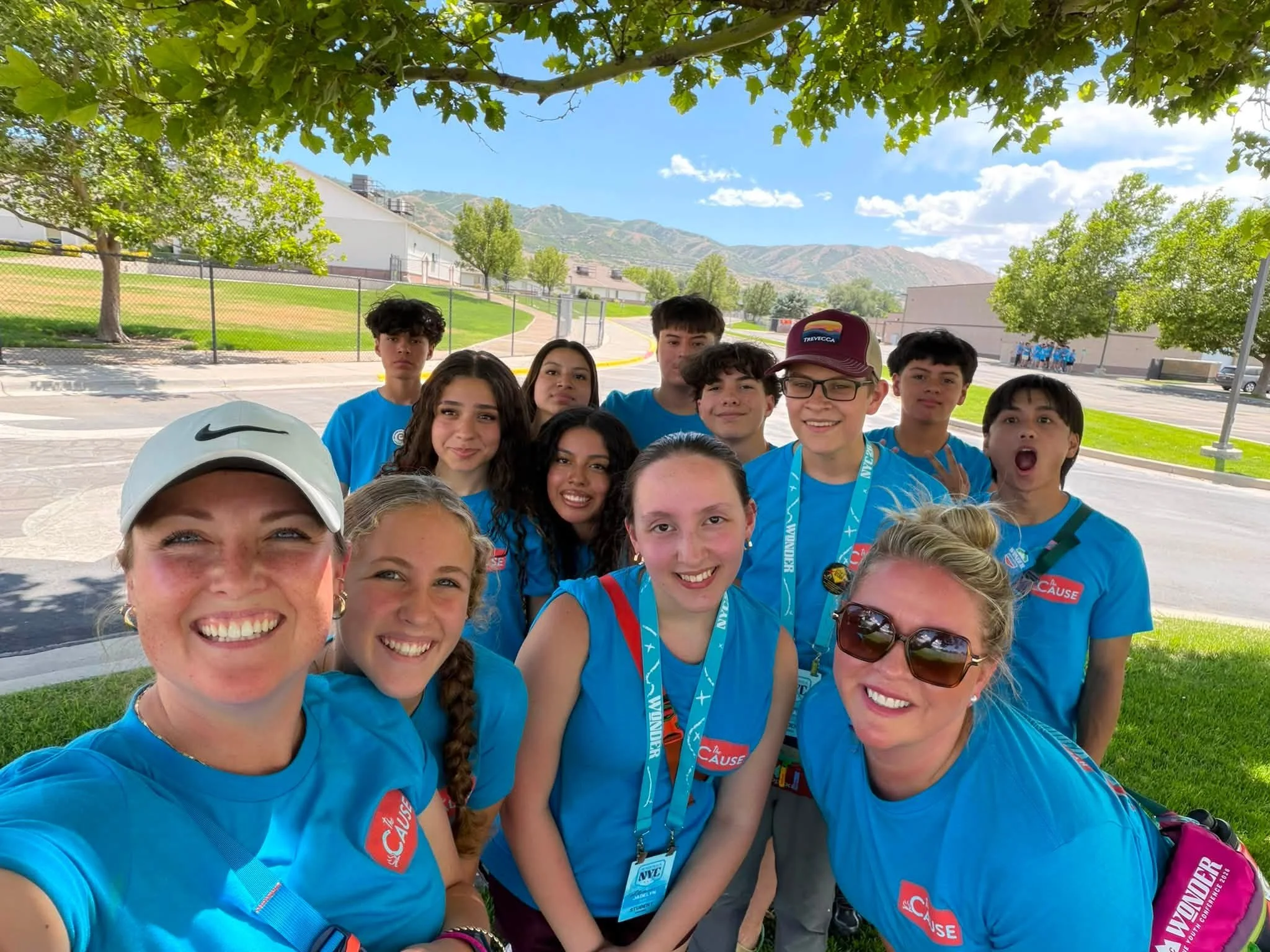 Group of smiling young people wearing blue shirts and event lanyards posing for a selfie outdoors under a tree with a school or community center in the background