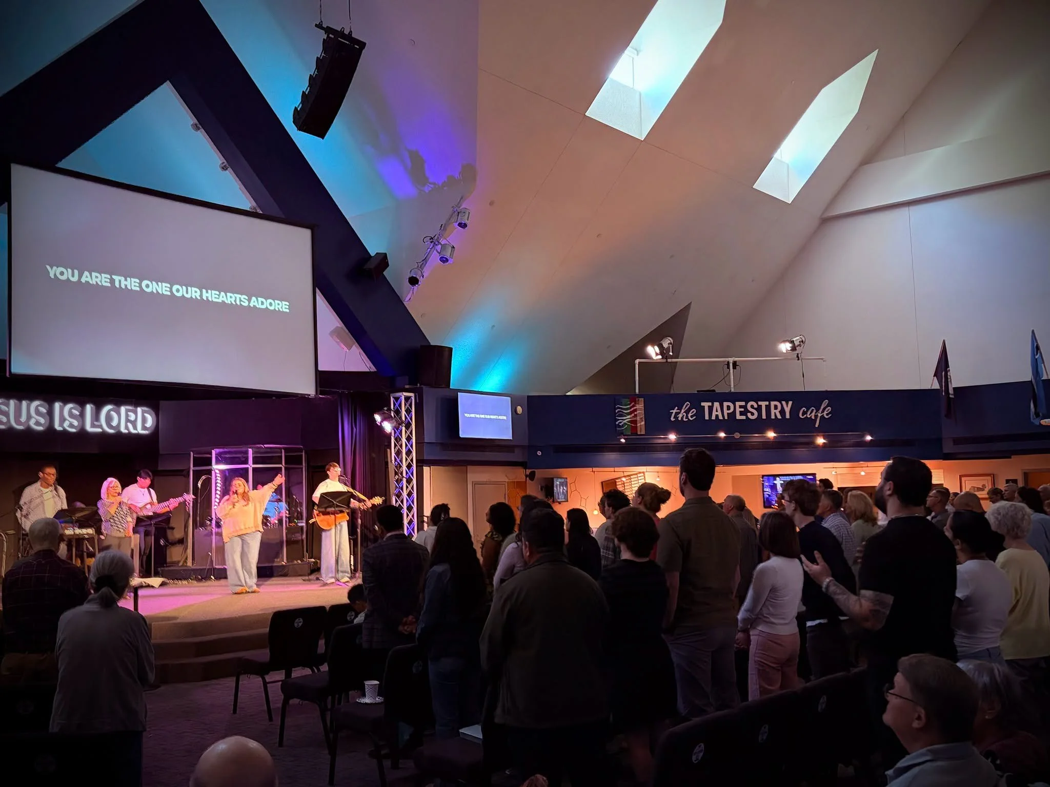 A church or community event with a stage where a group of people are singing or speaking, and the audience is standing and facing the stage. A large screen displays the text, 'YOU ARE THE ONE OUR HEARTS ADORE.' The venue has high ceilings with skylights and stage lighting.