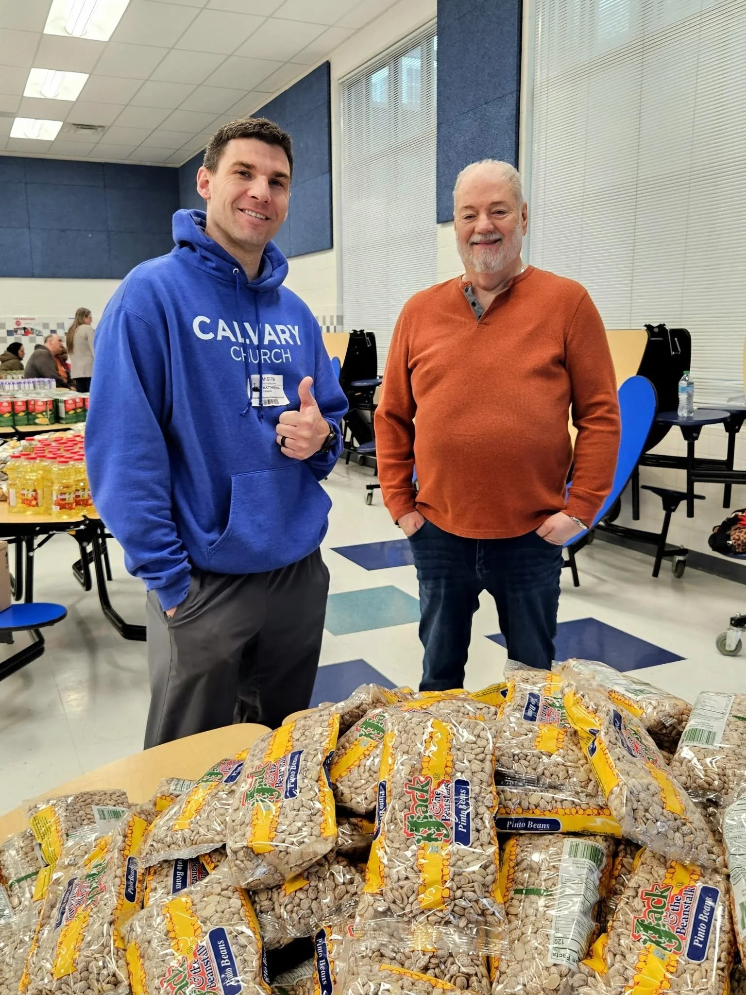 Two smiling men standing behind a table filled with bags of pinto beans in a community setting, with other people and tables with supplies in the background.