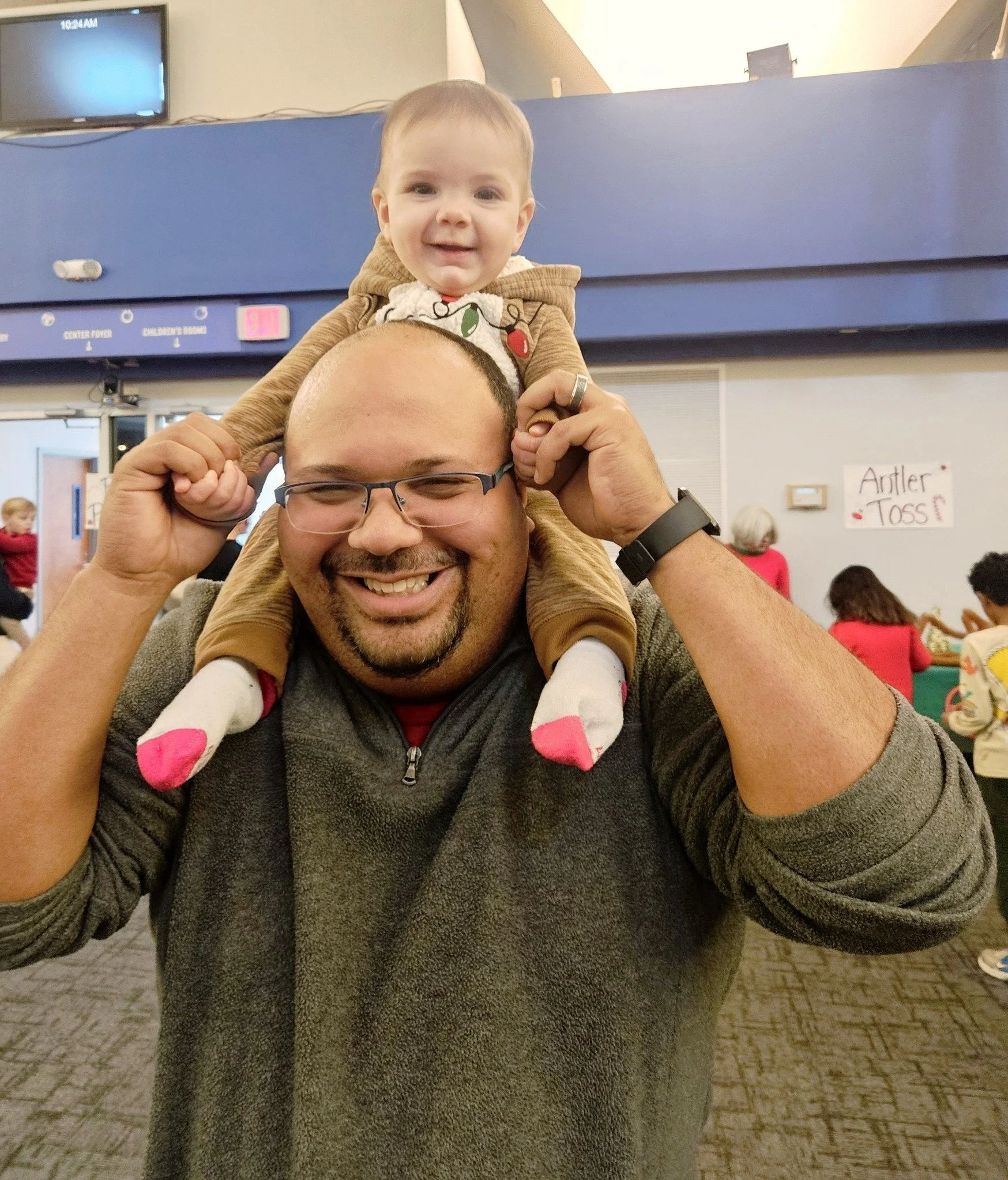 A man smiling while sitting with a young child on his shoulders in a community or recreational center, with other children and adults in the background.