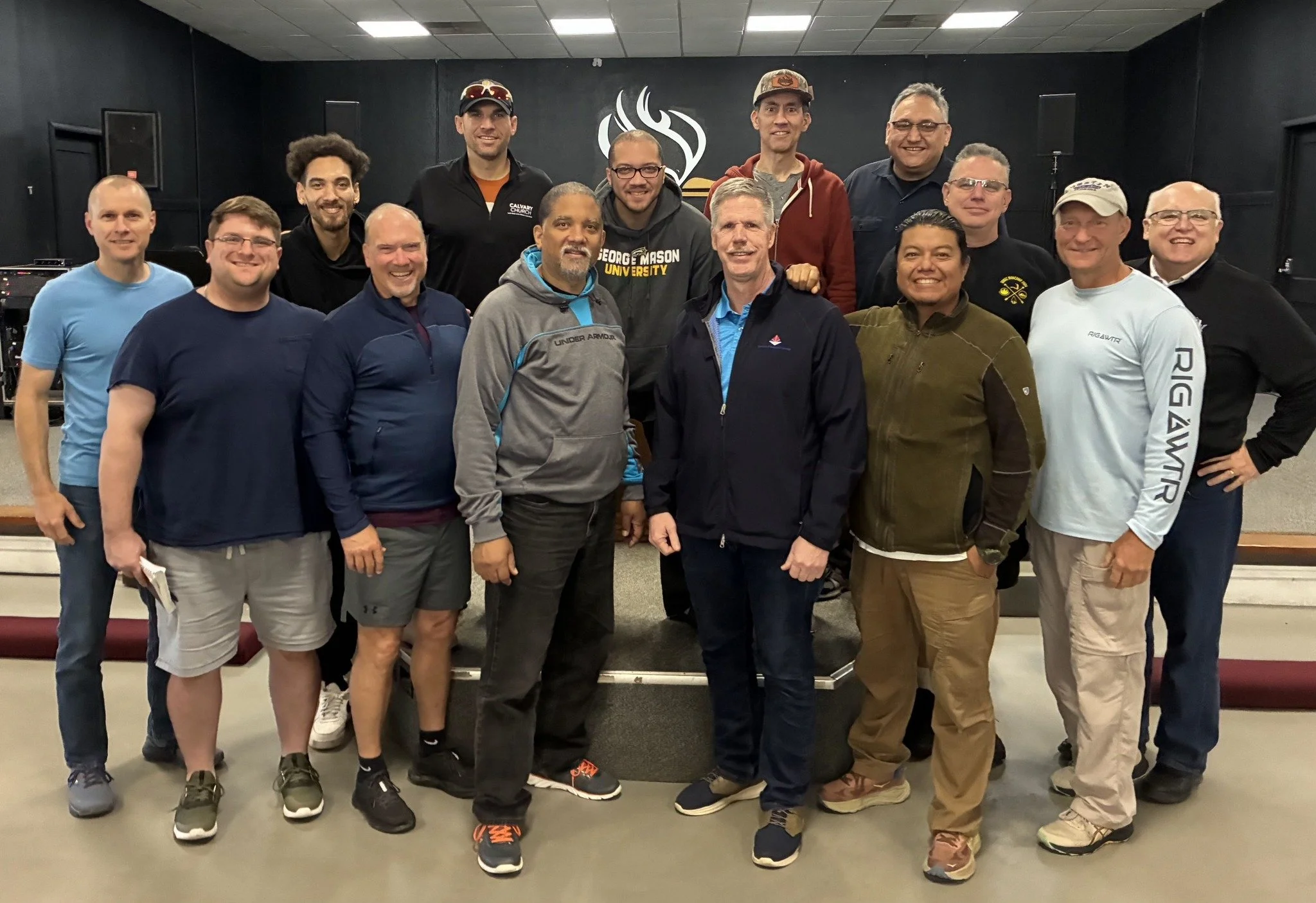 Group of fifteen men and women posing for a photo in a room with black walls and a logo in the background. They are smiling and standing close together.