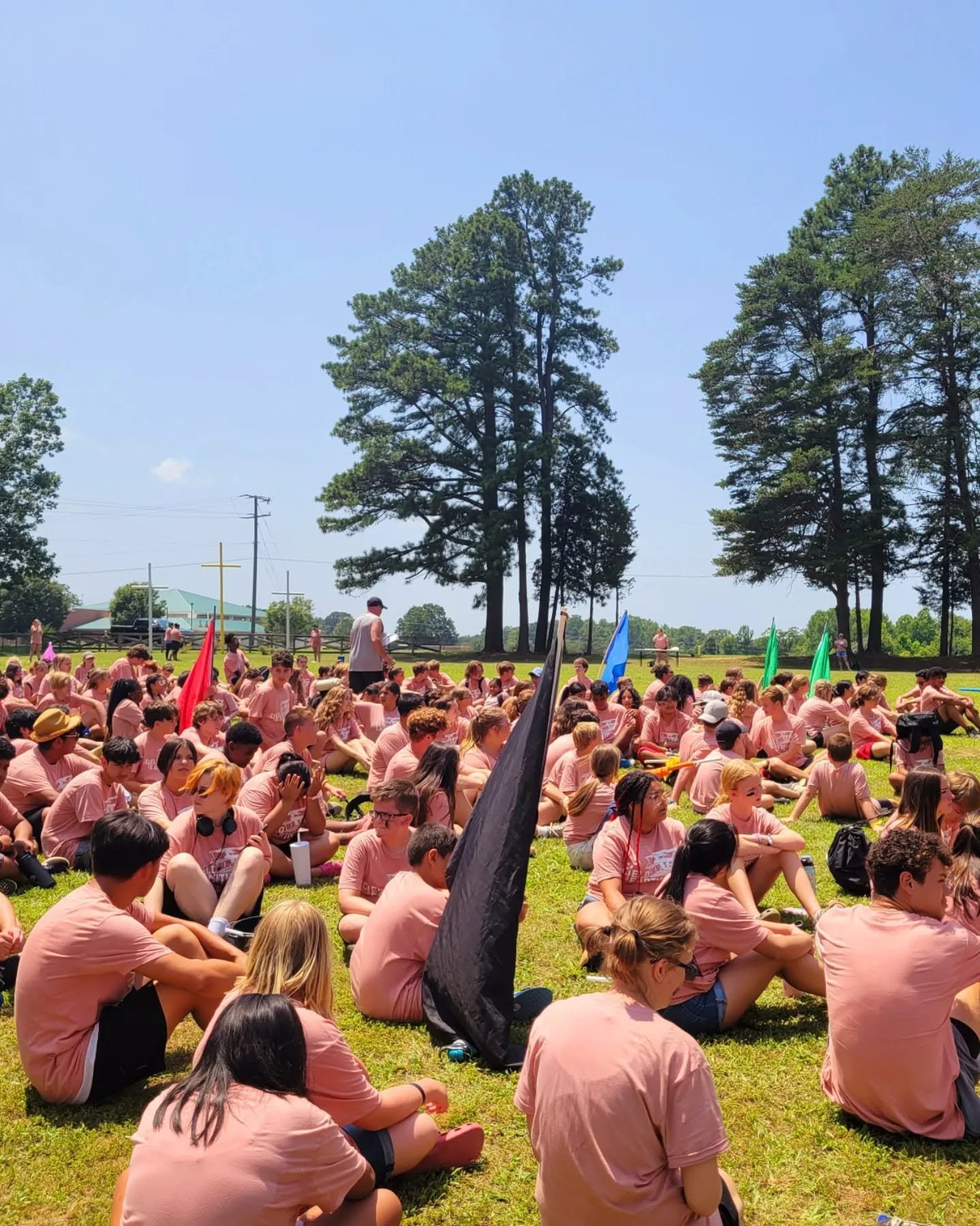A large group of people, mostly teenagers, sitting on the grass outdoors during daytime, wearing matching pink shirts, with some holding flags, under tall trees, with a blue sky overhead.