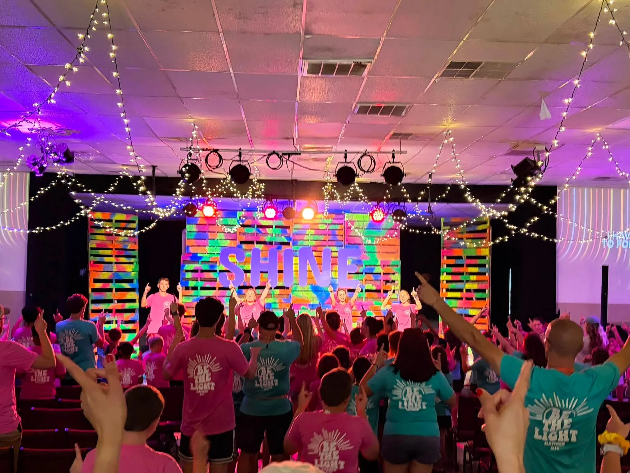 A large indoor gathering with many children and adults wearing pink and teal T-shirts, celebrating with hands raised. The stage is decorated with colorful 'SHINE' and 'BRIGHT' signs, and the ceiling is adorned with string lights and hanging stage lights.