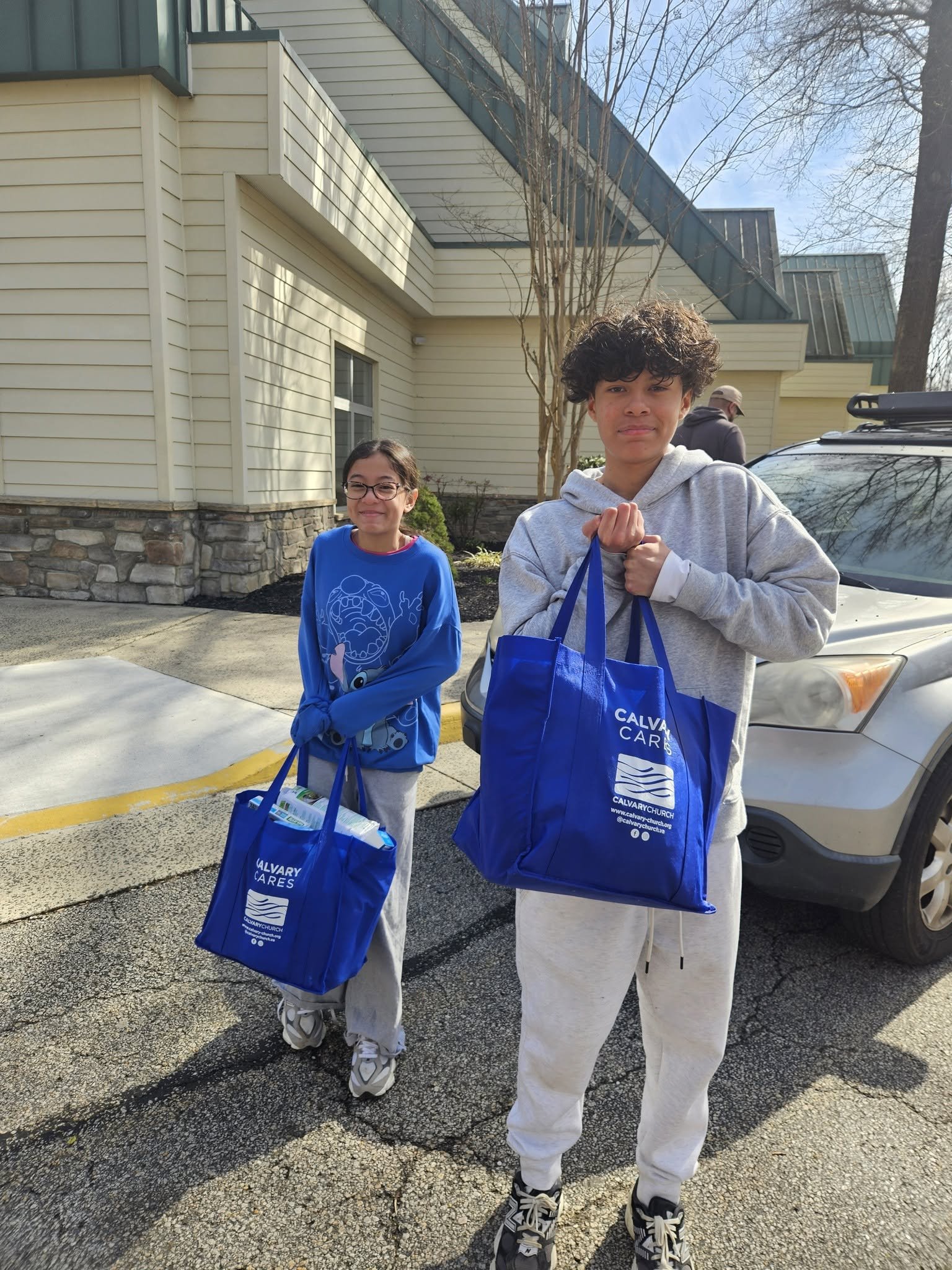 Two young people holding shopping bags outside near a parked car. They are smiling and standing in front of a beige building with stone foundation. The sky is clear and there are leafless trees in the background.