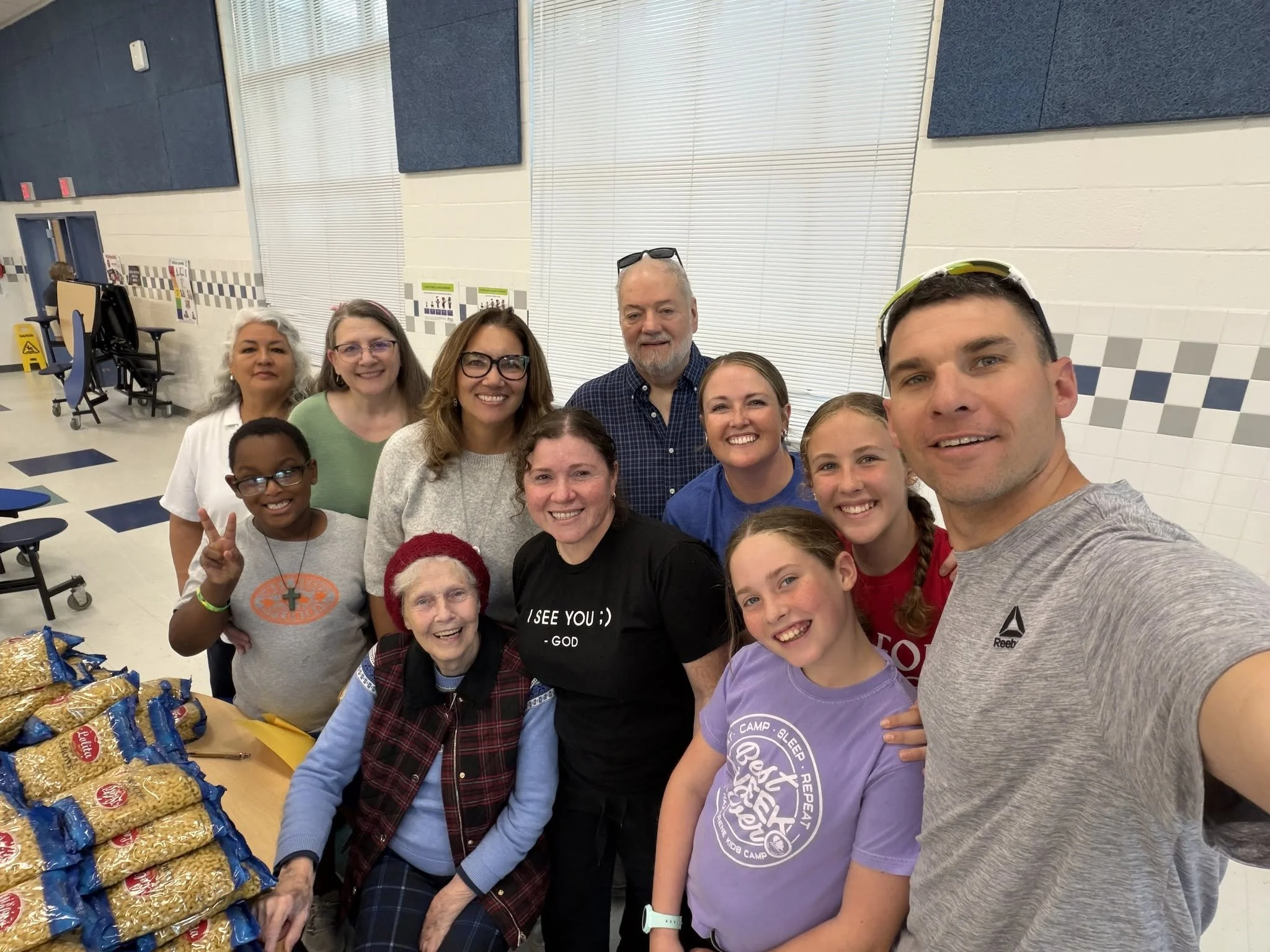 Group of smiling people in a room with blue and white walls, some wearing glasses and casual clothing, and a table with food packets in front of them.
