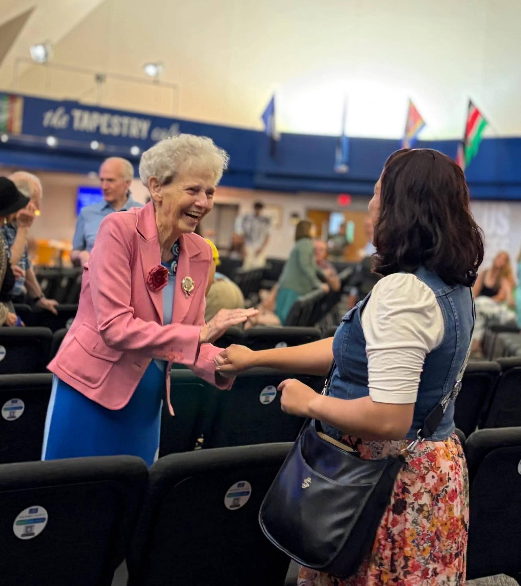 Two women shake hands in a crowded indoor space, smiling at each other. One woman has gray hair and is wearing a pink blazer, while the other has dark hair and is wearing a denim vest and floral skirt.