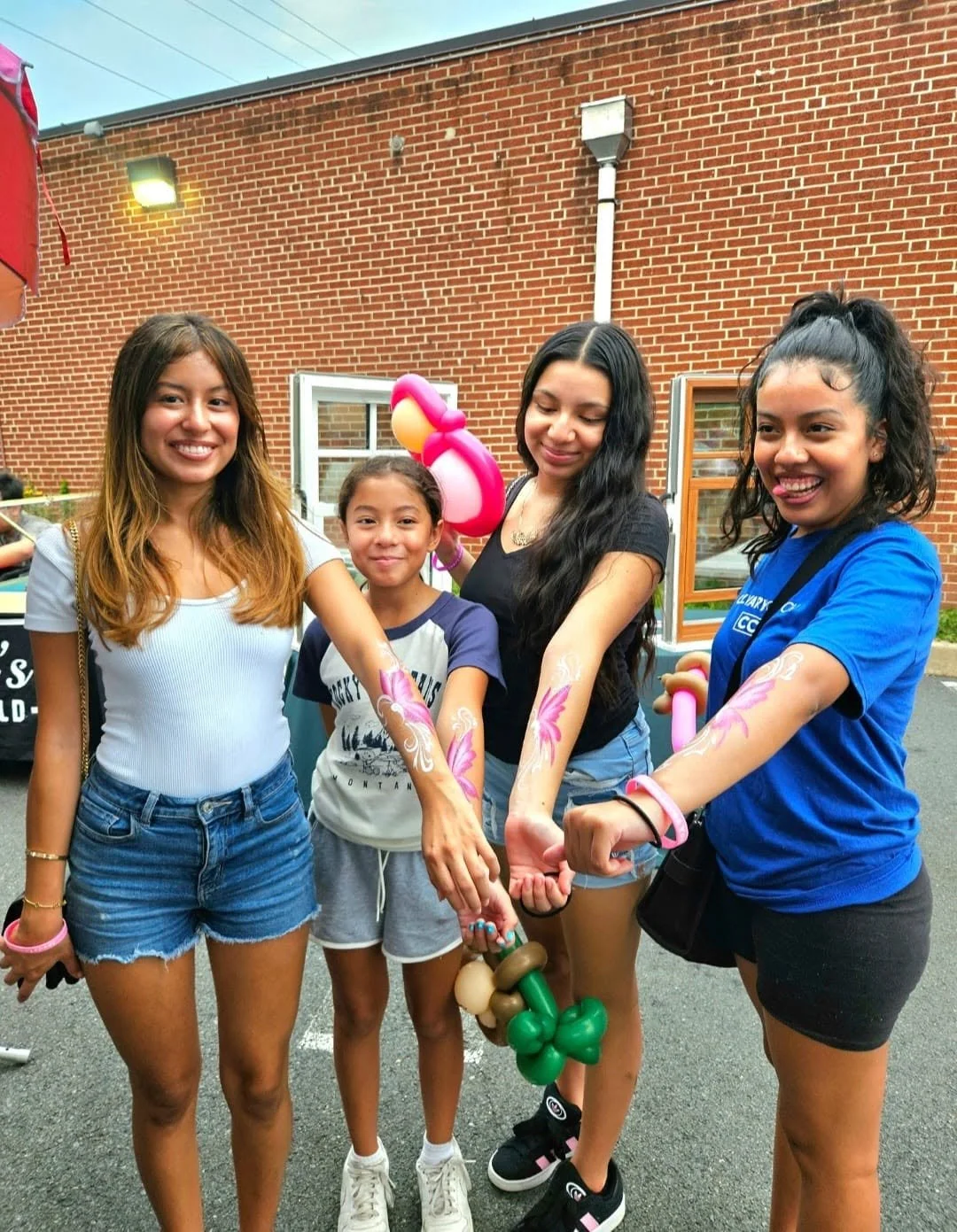 Group of four girls with face paint and balloon art, standing outdoors with a brick building background, smiling and showing their painted arms.