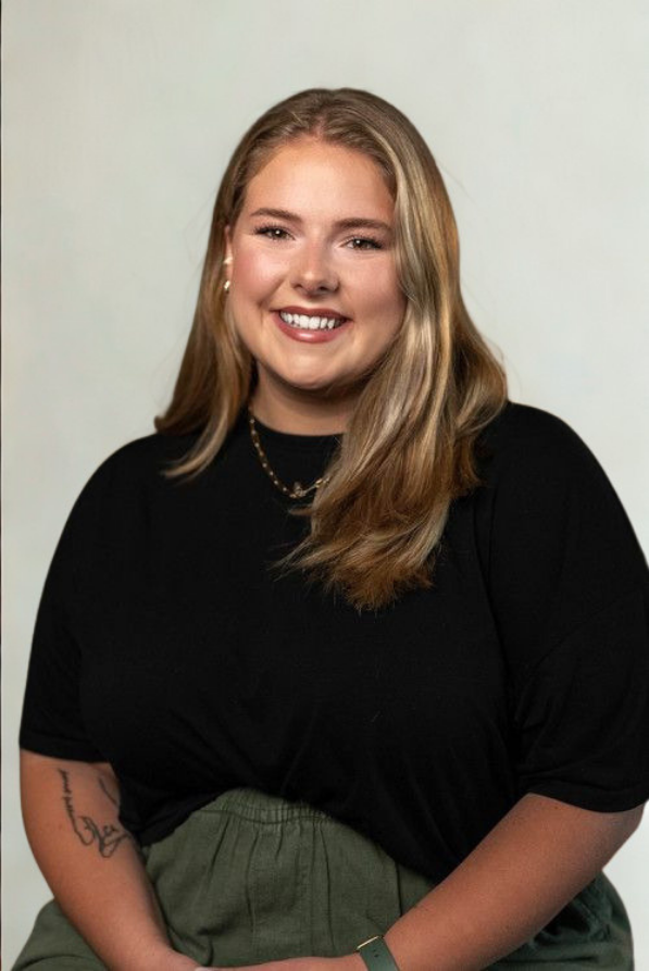A smiling woman with shoulder-length blonde hair, wearing a black top, green pants, and gold jewelry, posed against a plain light-colored background.