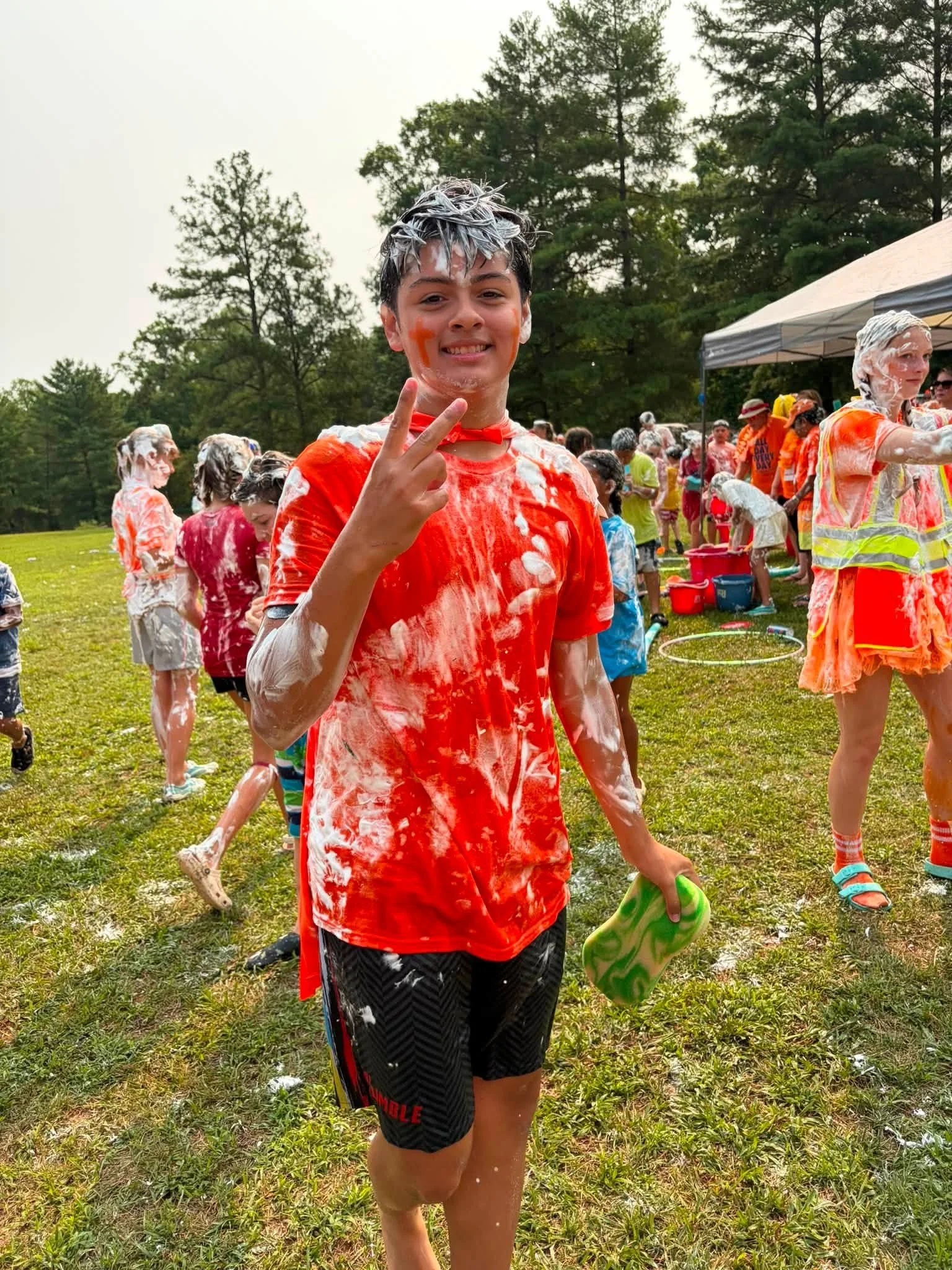 Young boy with face paint and soap foam hair, smiling and making a peace sign at an outdoor foam party, with other participants and a canopy tent in the background.