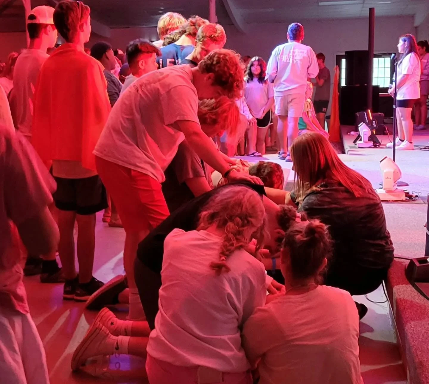 People gathered in a room, some sitting on the floor and others standing, engaging in a first aid training or CPR class with a focus on a person lying on the ground, illuminated by red lighting.