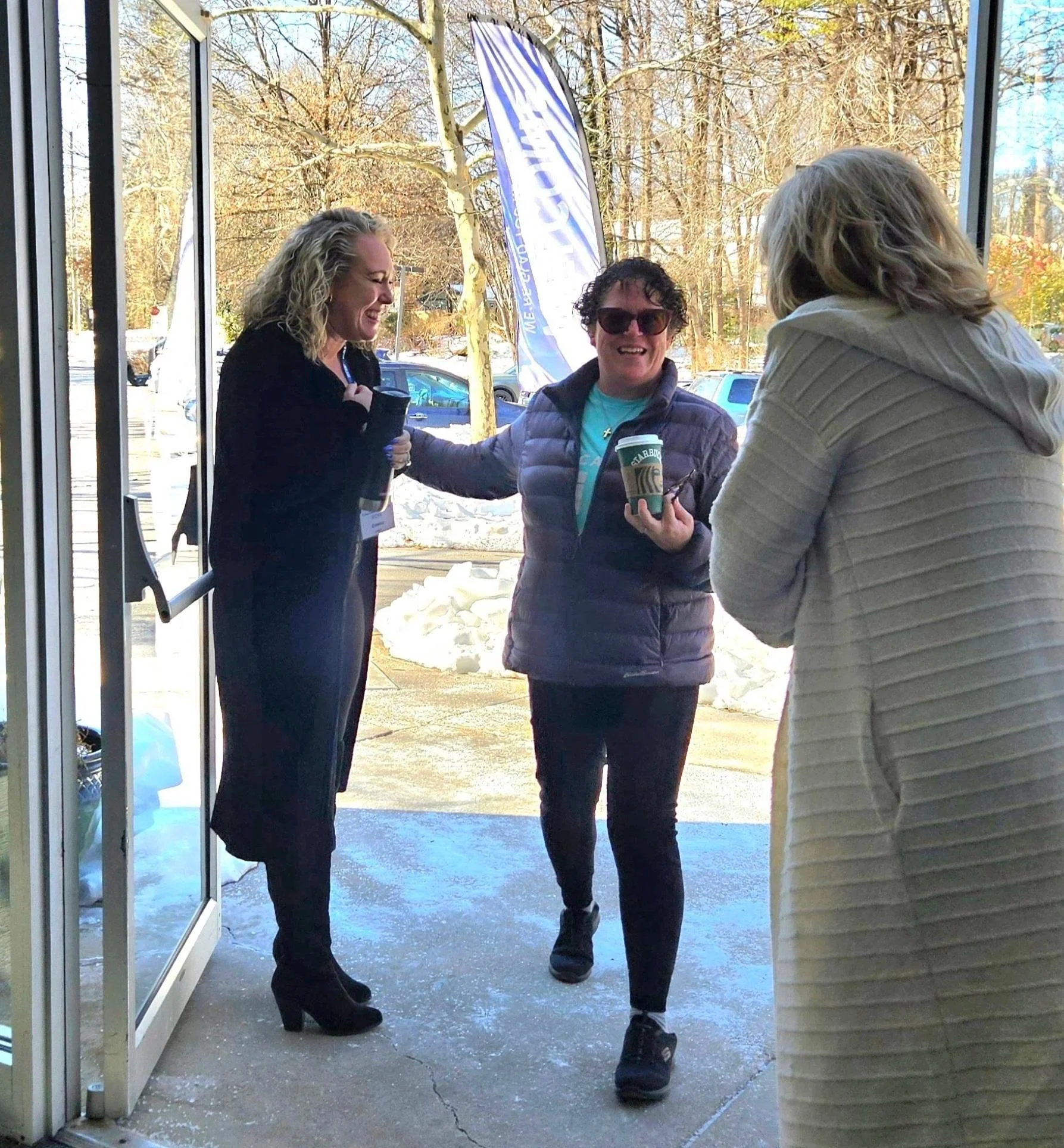 Three women standing outside the entrance of a building, chatting and smiling, with snow on the ground and trees in the background.