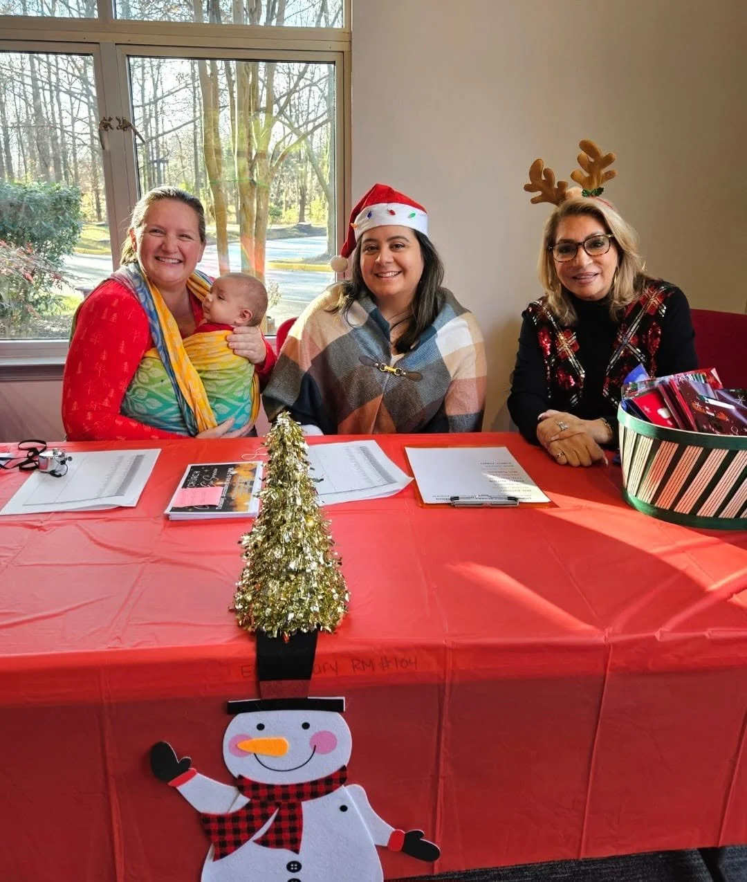 Three women sitting at a red Christmas table with papers and a small gold Christmas tree, celebrating holiday season. The woman on the left is holding a baby, the woman in the center wears a Santa hat, and the woman on the right has reindeer antlers. A snowman decoration hangs on the front of the table.