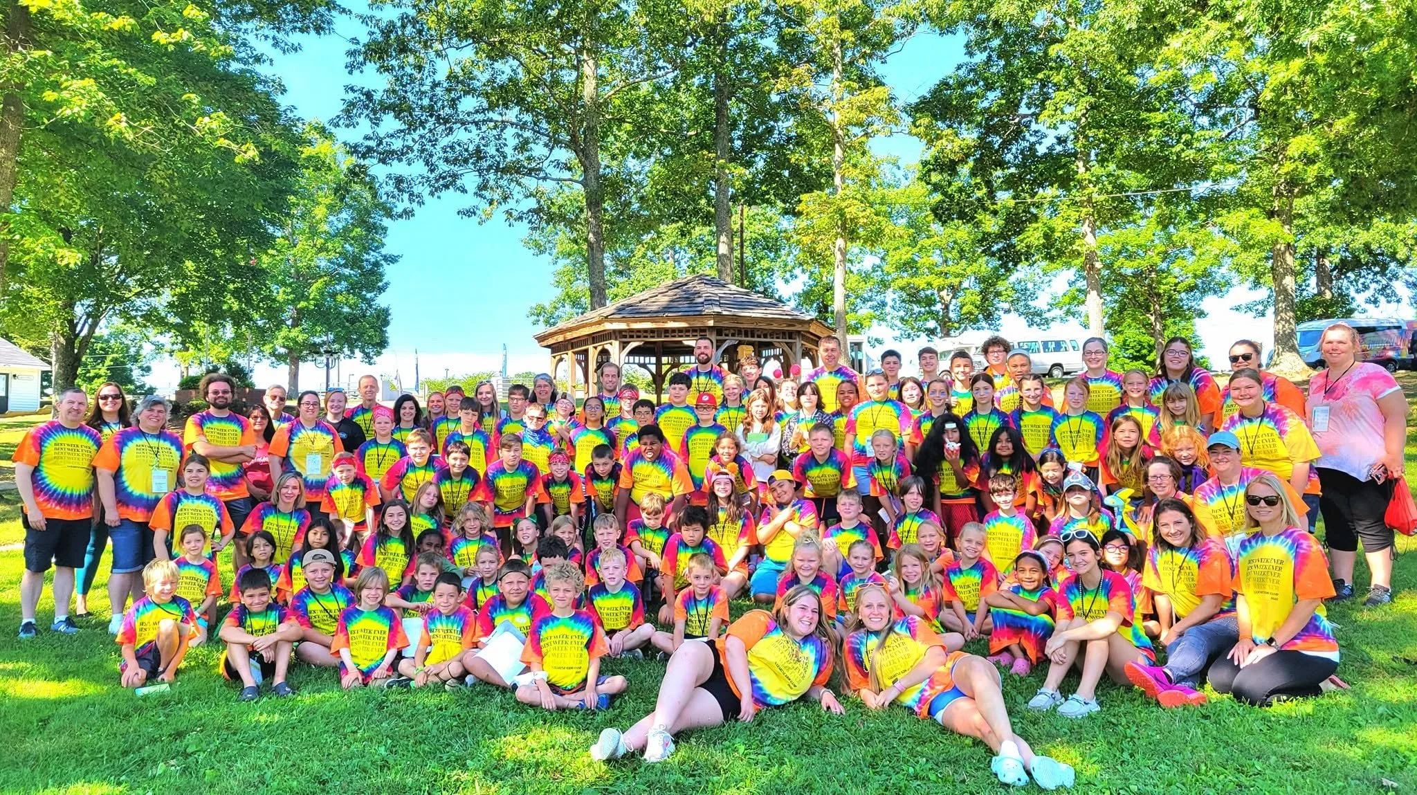 Large group of children and adults gathered outdoors under trees, many wearing matching tie-dye T-shirts with yellow text, in front of a gazebo on a sunny day.