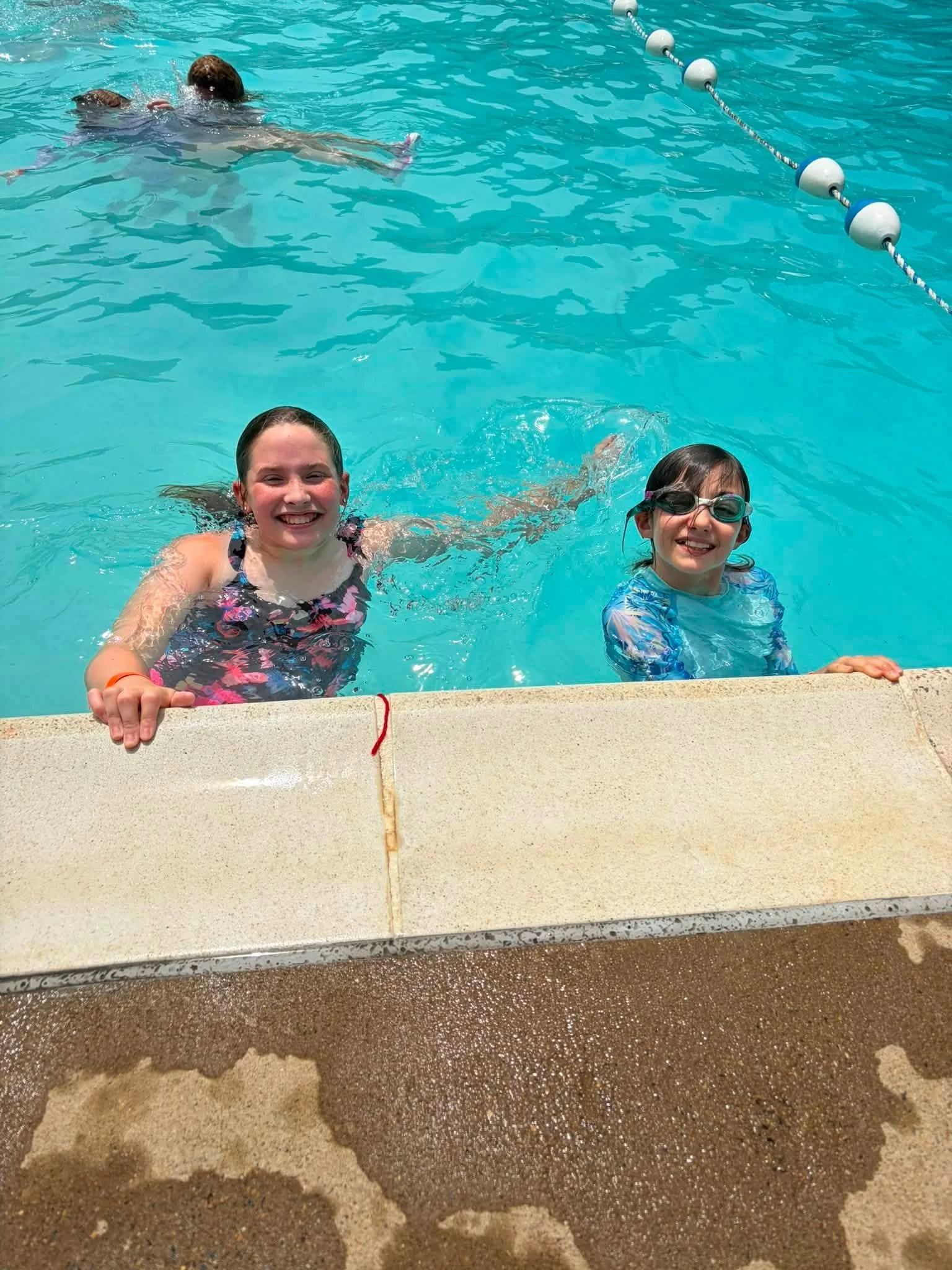 Two young girls smiling and enjoying themselves in a swimming pool, wearing swimsuits and goggles, near the edge of the pool with a lane marker visible in the background.