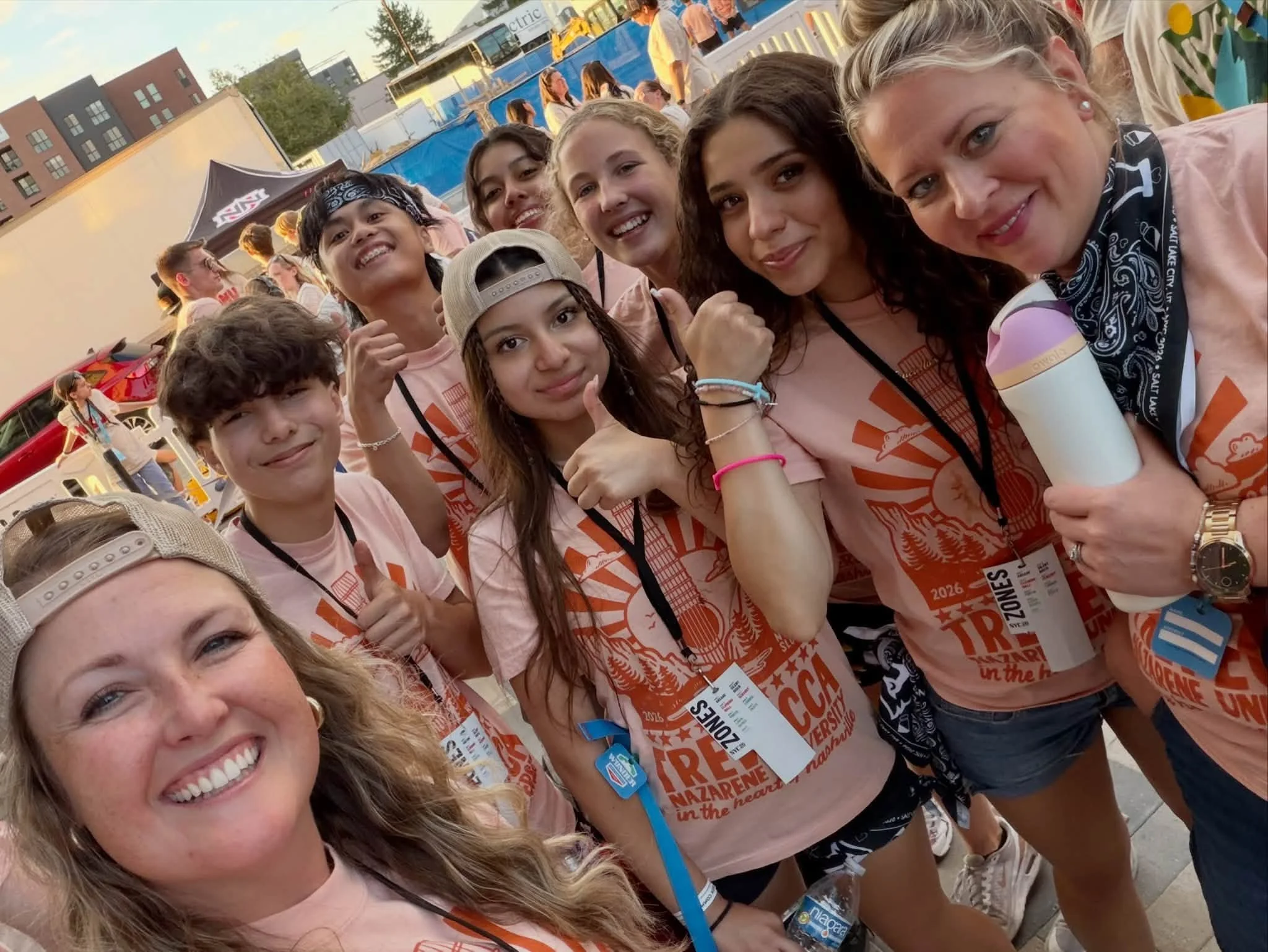 Group of smiling young women and girls at an outdoor event, wearing matching pink T-shirts with a design and identification badges, with some making fists or thumbs-up gestures.