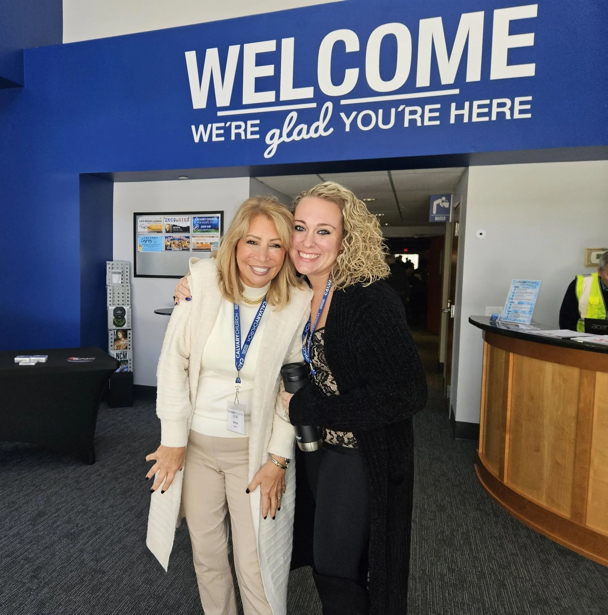 Two women smiling and posing for a photo at an indoor event, standing under a blue sign that reads 'Welcome, We're glad you're here'; both women wear conference badges.