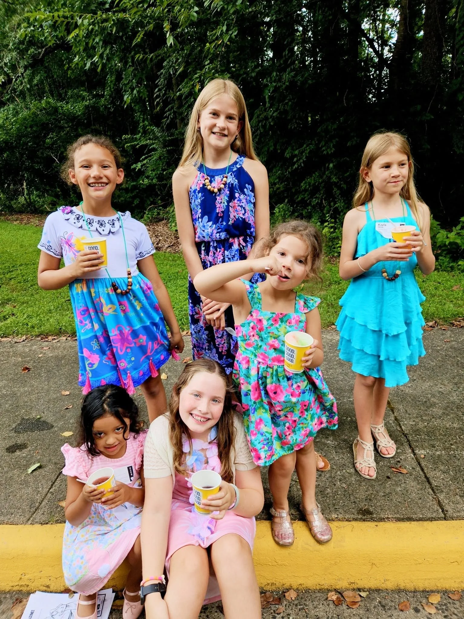 Group of young girls outdoors, some holding cups, smiling and standing on a sidewalk with greenery in the background.