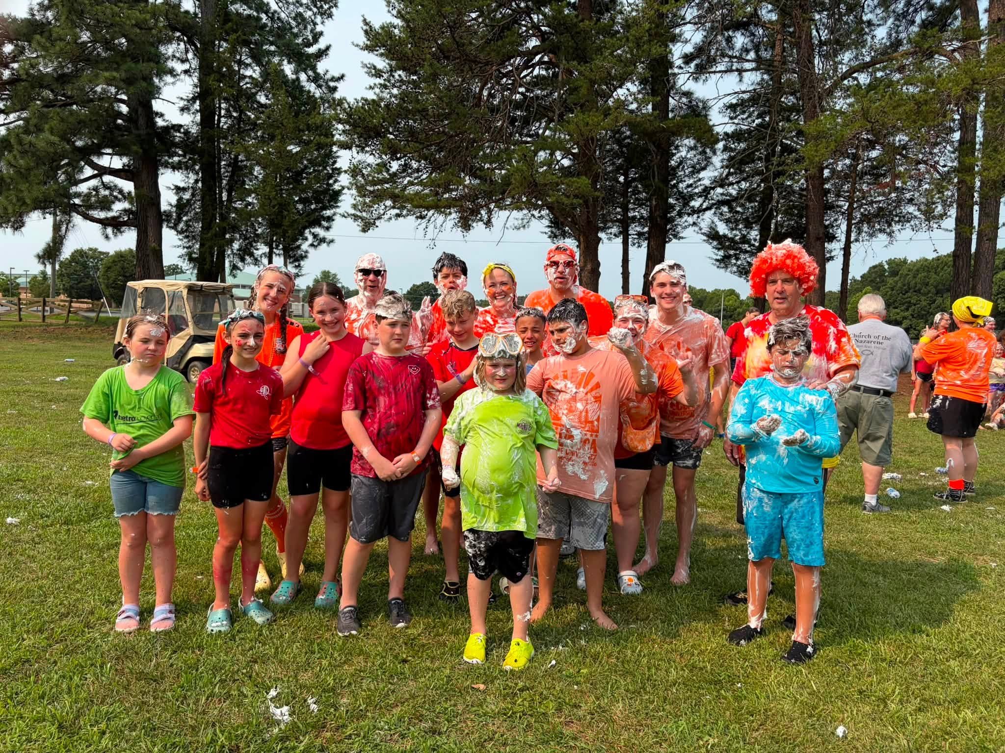 A group of people, including children and adults, gathered outdoors on grass, covered in foam and wearing colorful clothes, with trees in the background.
