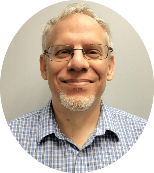 A middle-aged man with glasses and a white beard, smiling, wearing a blue checkered dress shirt, standing against a light gray background.