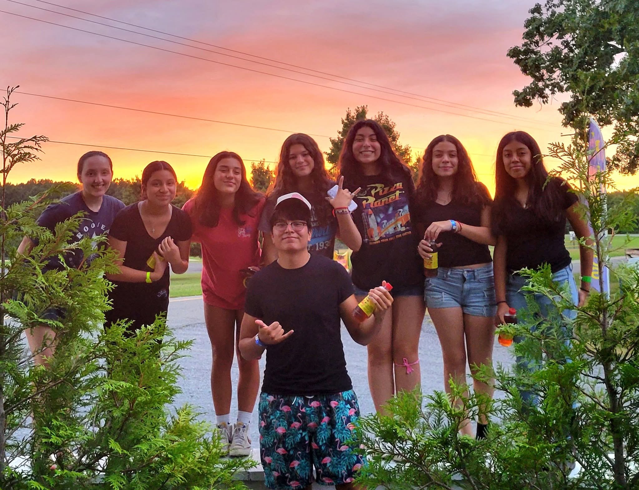 A group of people standing outdoors during sunset with colorful sky in the background, surrounded by green plants and trees.