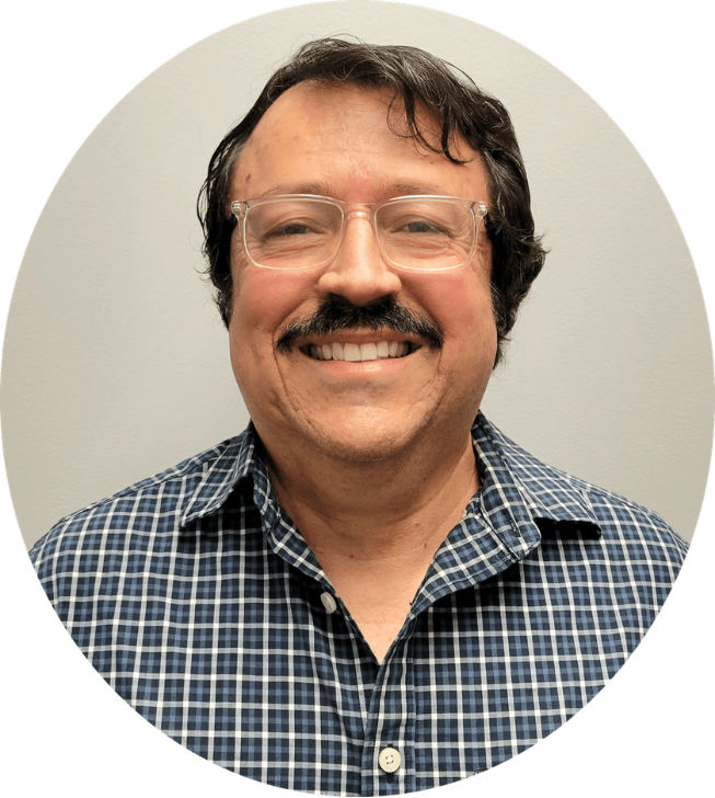 Smiling man with glasses and mustache wearing a checkered shirt, standing against a plain light-colored wall.