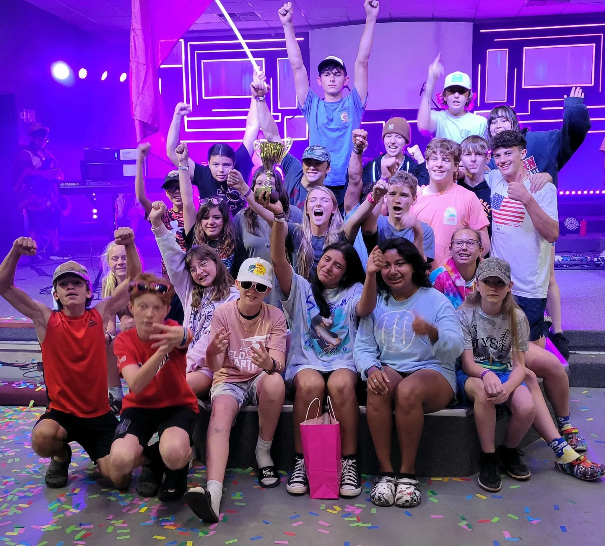 A group of children and teenagers at a celebration event, some holding up arms and a trophy, with colorful confetti on the floor and purple neon light backgrounds.