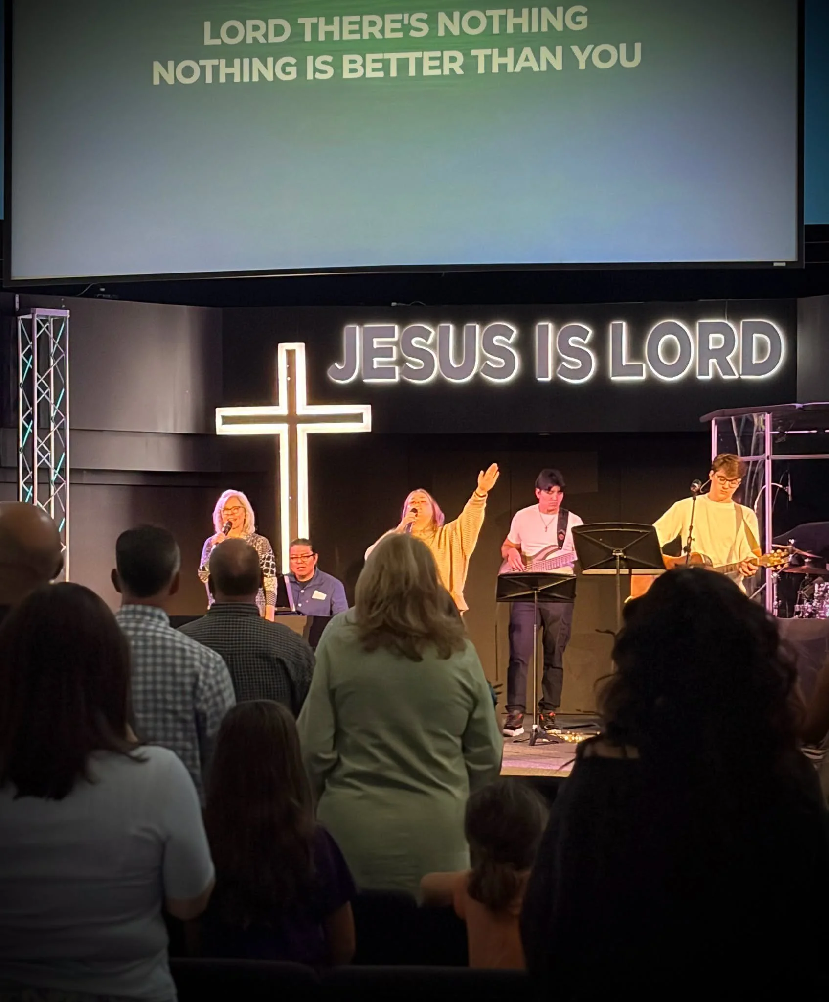 A church service or gathering with a cross and illuminated sign reading 'JESUS IS LORD'. A large screen displays the message: 'LORD THERE'S NOTHING, NOTHING IS BETTER THAN YOU'. People are standing and some praying or singing, with many facing the stage.
