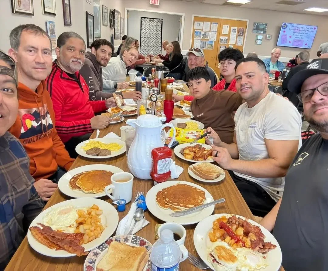 People sitting around a long table in a restaurant, having breakfast with plates of pancakes, bacon, eggs, toast, and other breakfast foods.