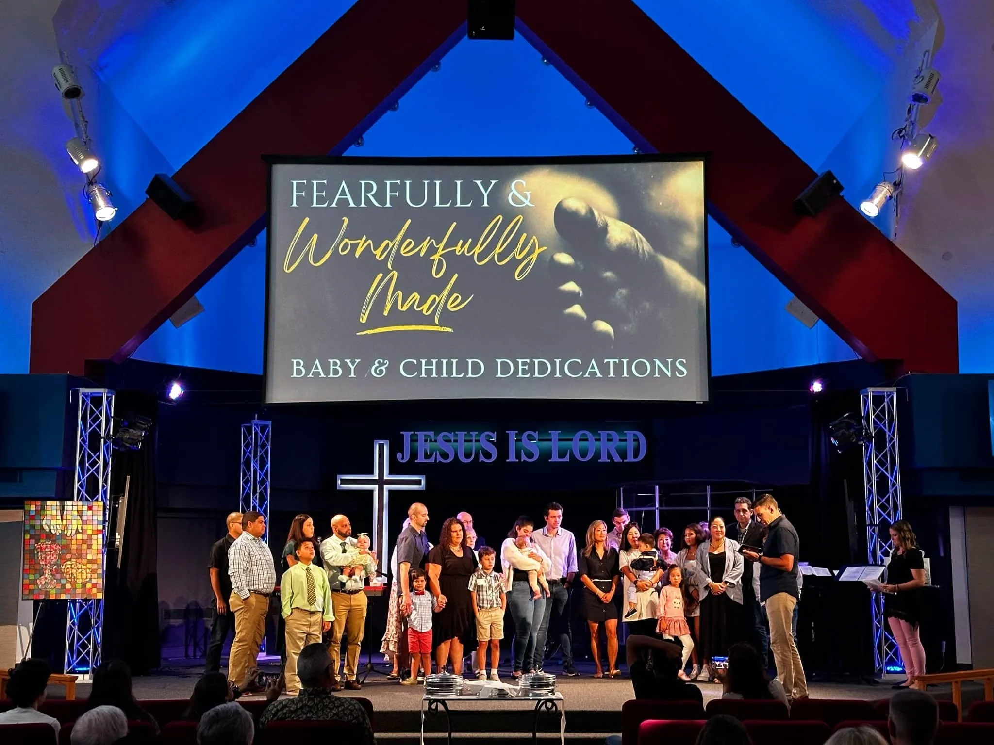 A large group of people gathered on stage for a baby and child dedication ceremony with a sign behind them that reads 'Fearfully & Wonderfully Made' and a space-themed background. Audience members are seated in front.