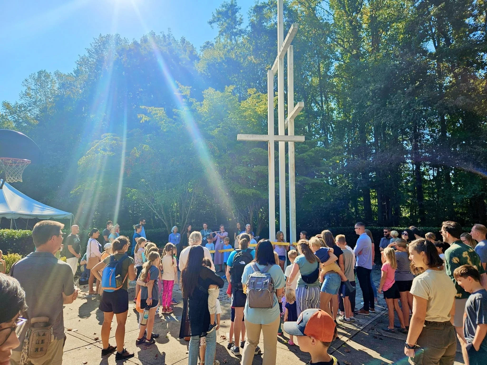 A crowd of people gathered outdoors on a sunny day with trees in the background. There is a large cross structure in the center and a basketball hoop on the left. The crowd appears to be listening or participating in an event.