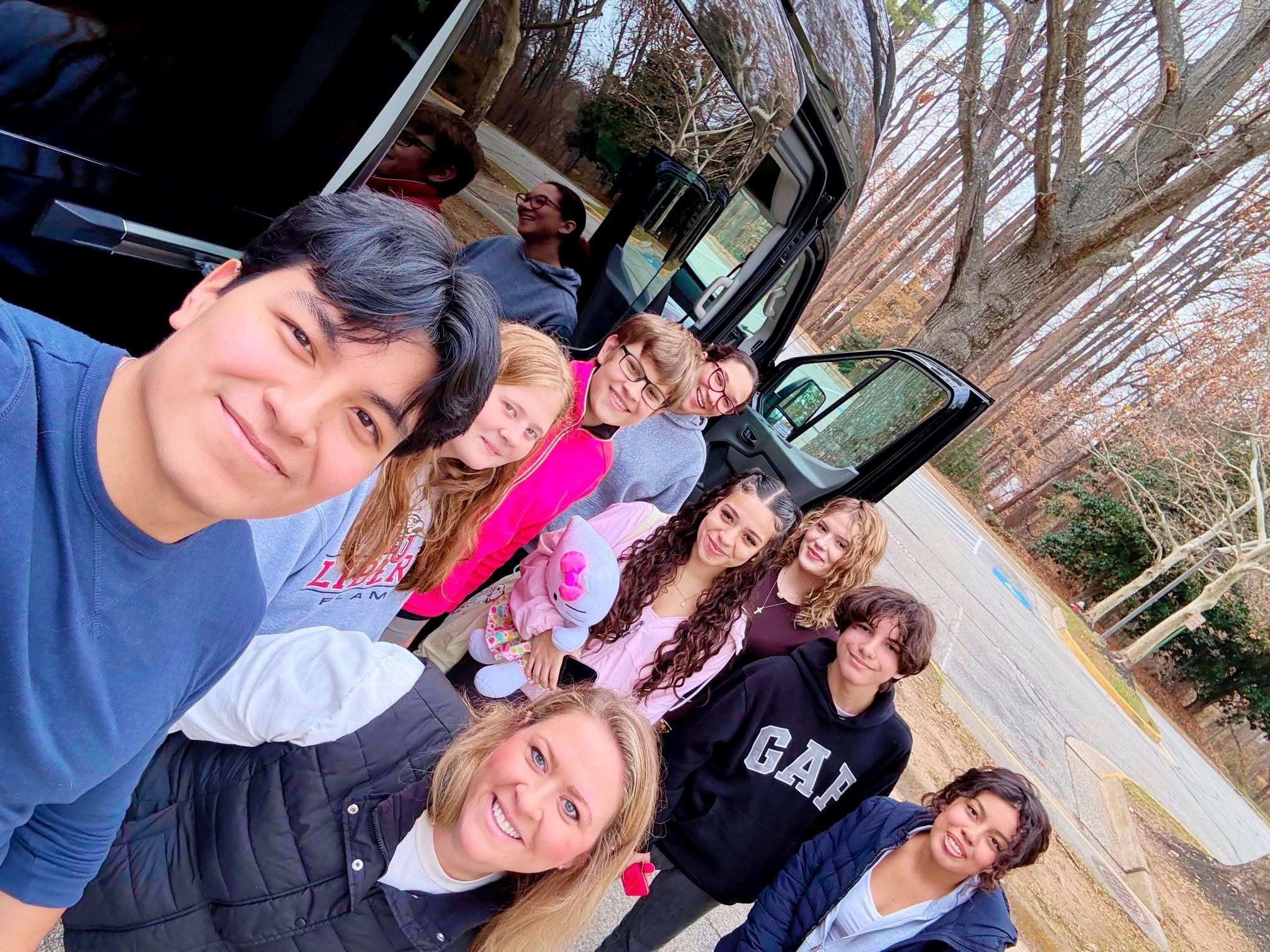 Group of people taking a selfie outdoors in a wooded area during autumn, with trees shedding leaves and a road in the background.