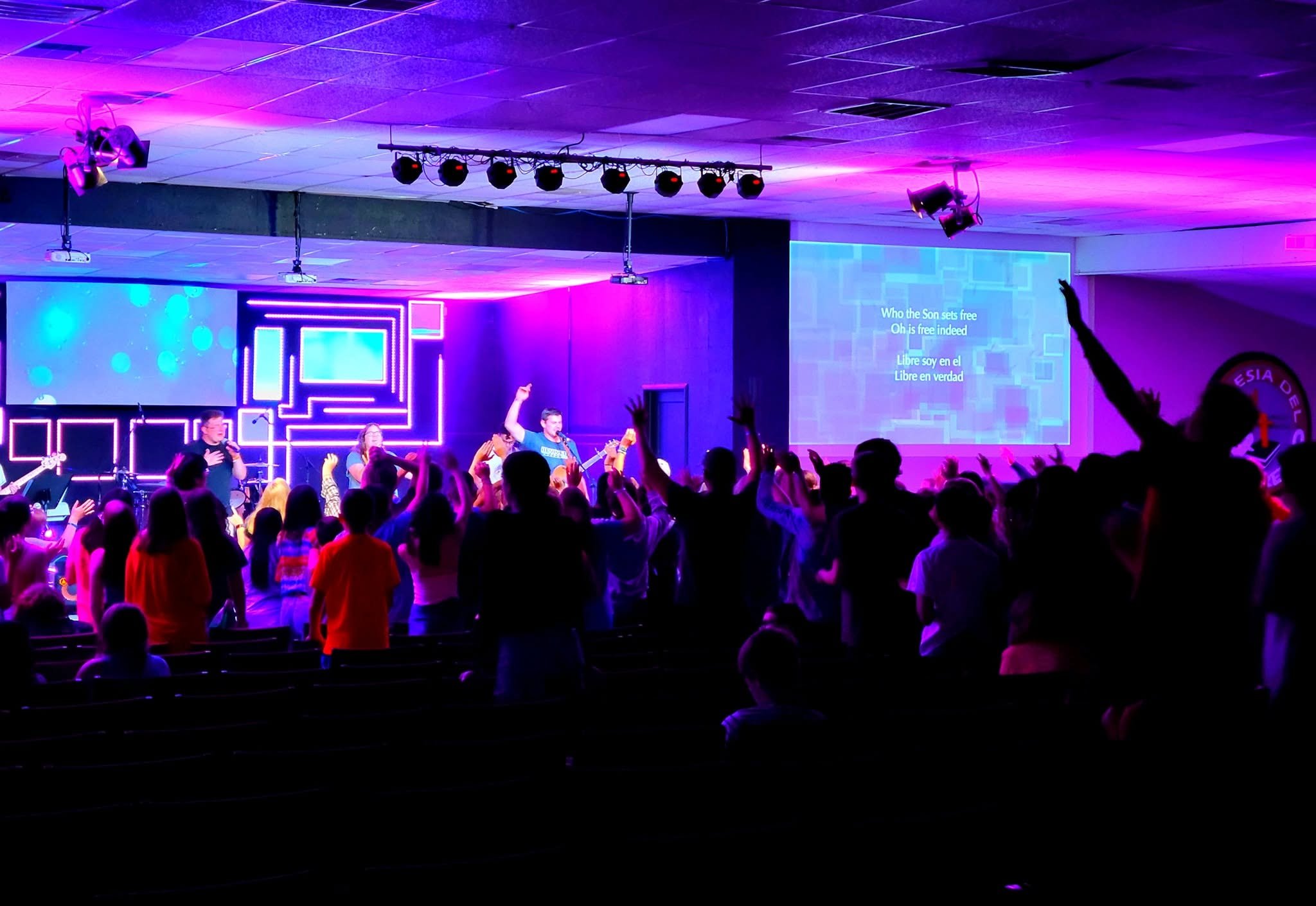 A large gathering of people in a room with colorful purple and blue lighting, participating in a musical or worship event with some raising their hands, a stage with musicians, and song lyrics projected on a screen.