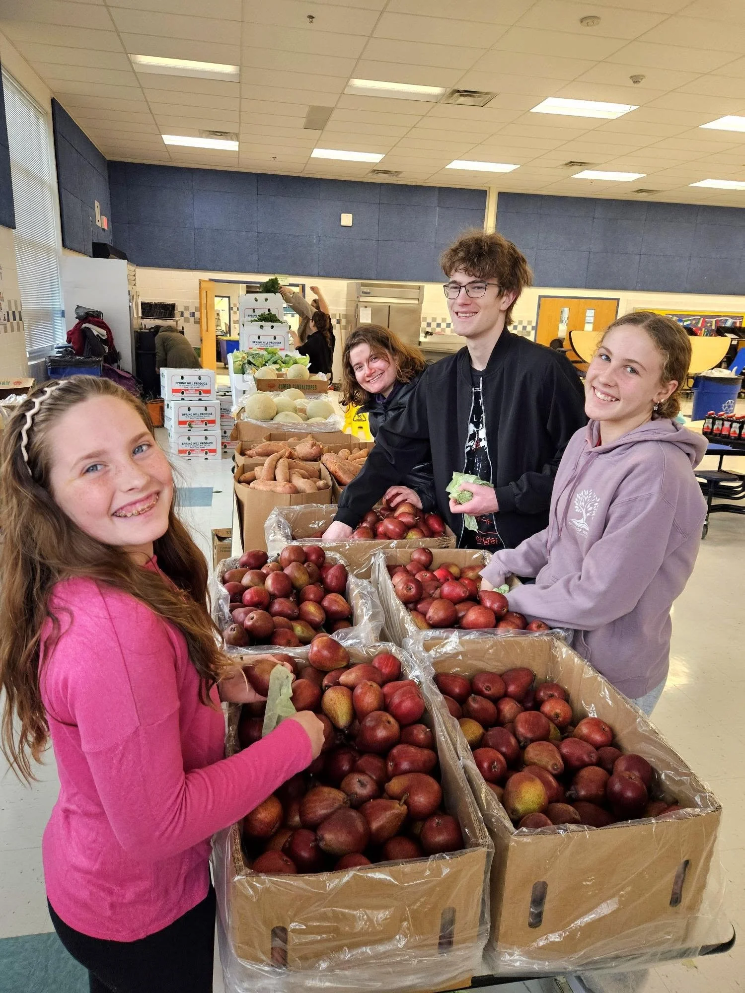 Group of people at a table with apples and sweet potatoes, inside a community center or cafeteria.