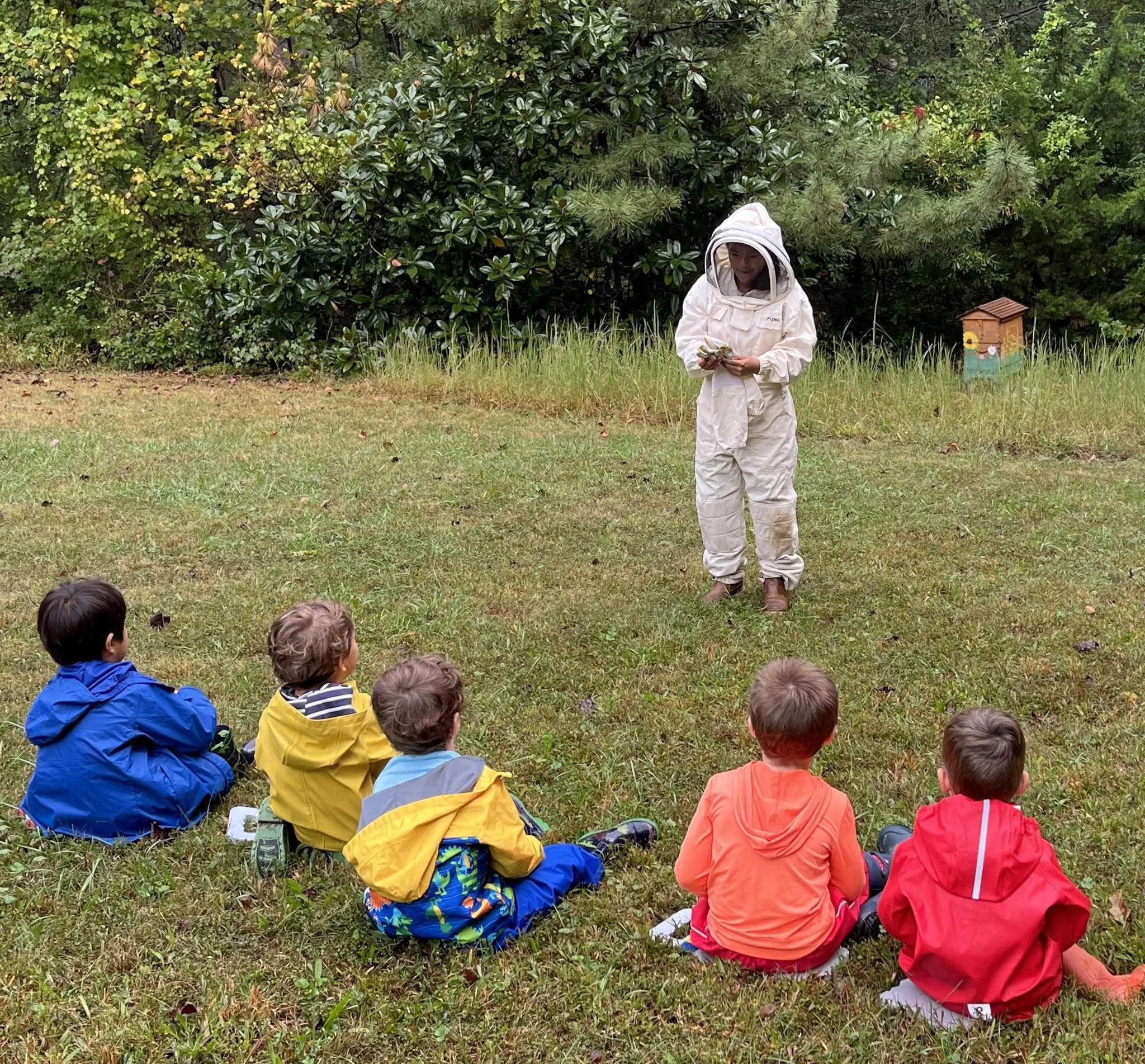 A group of children sitting on grass and listening to a woman in a beekeepers suit, who is holding a honeycomb, outdoors with greenery in the background.