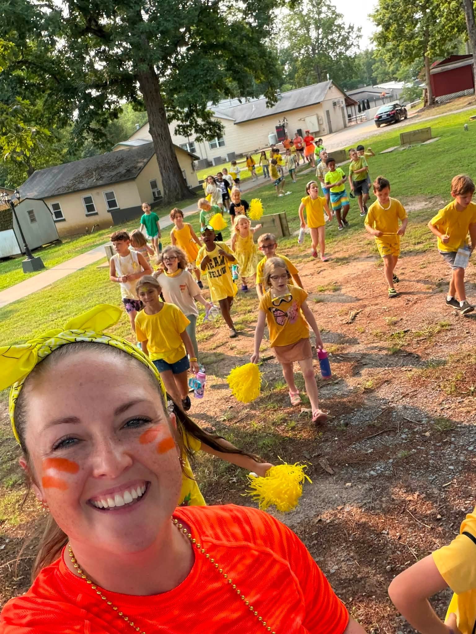 A woman with painted face smiling in the foreground, with children wearing colorful clothing and holding pom-poms in the background during an outdoor event on a sunny day.