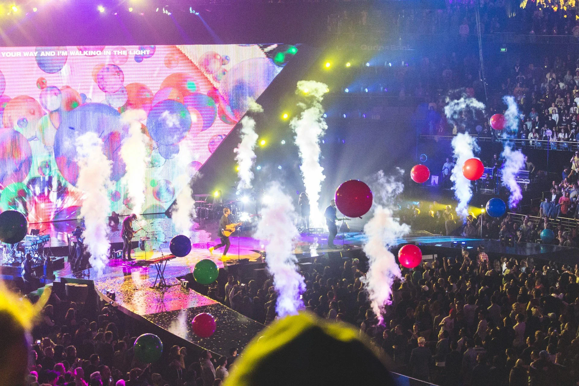 A concert with a stage, colorful lights, smoke effects, large red and green balloons, and a crowd of people watching a band perform.