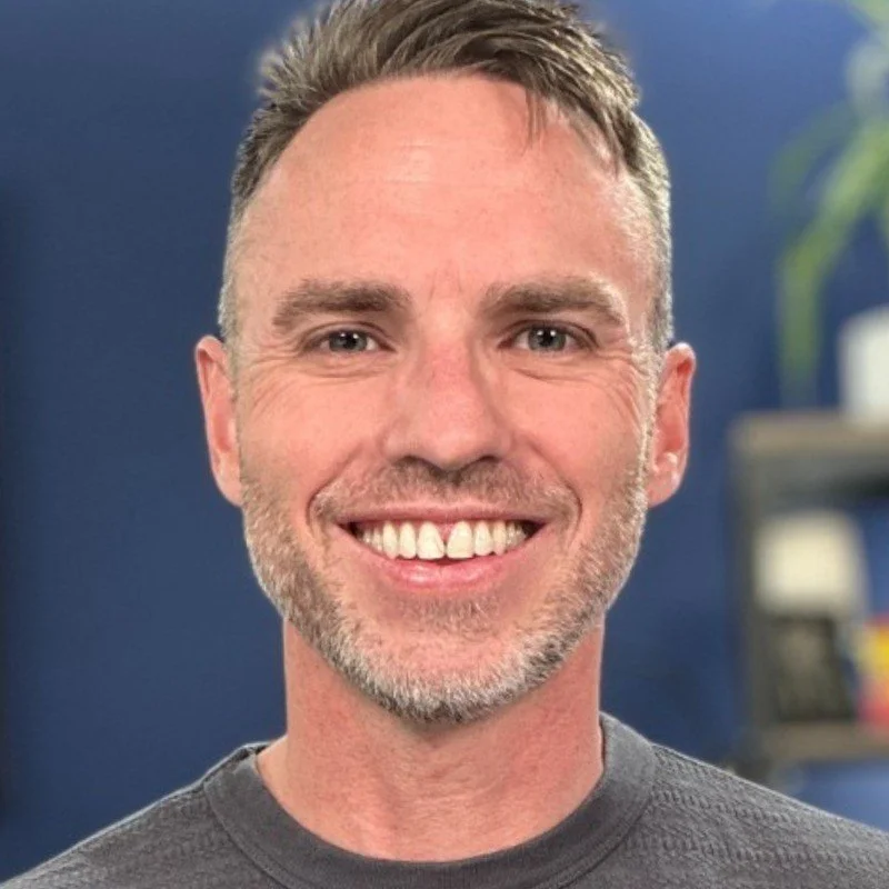 Close-up of a smiling man with short brown hair and a beard, wearing a dark grey shirt, indoors with a blue wall and blurred bookshelf in the background.