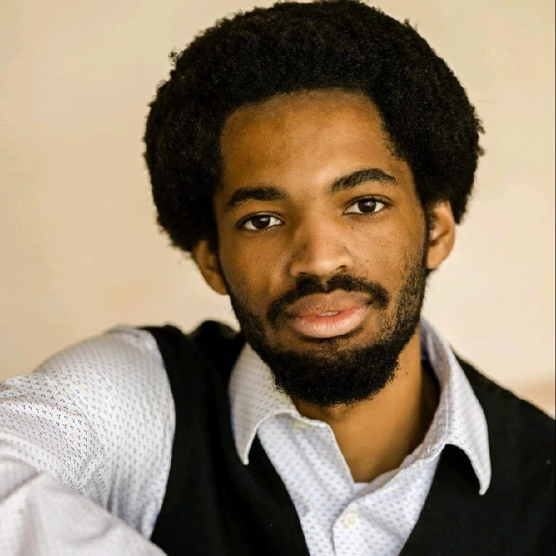 A young man with a beard and afro hairstyle wearing a white collared shirt and a black vest, looking directly at the camera.