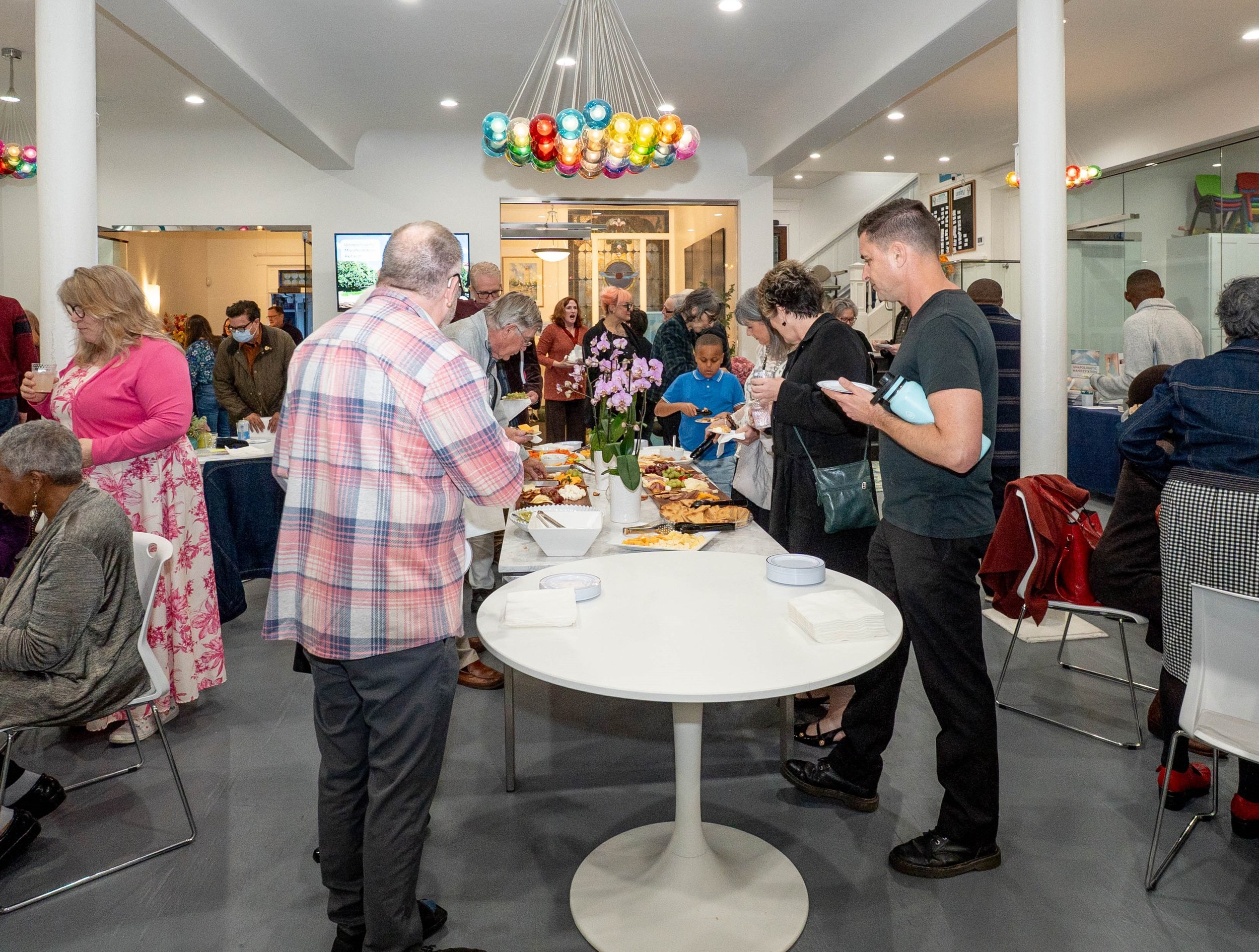 People gathering at a buffet table with food and flowers, inside a well-lit room with colorful hanging lights.