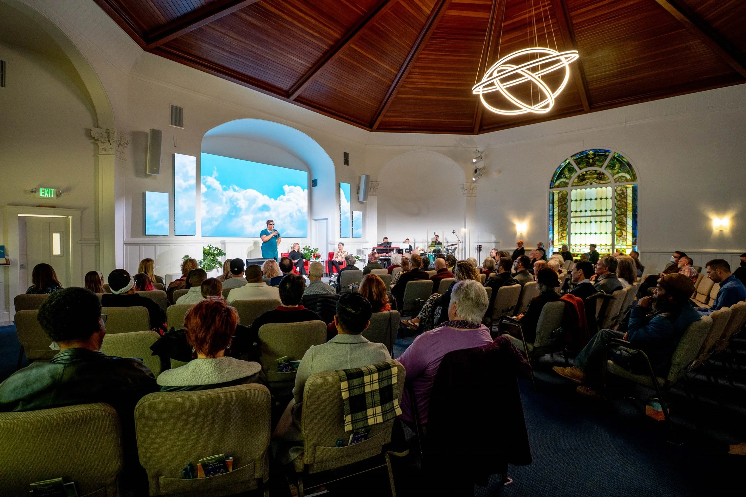 An indoor event hall filled with seated audience listening to a speaker on stage. The stage has a large screen displaying a sky with clouds, and there is a band setup. The hall features stained glass windows, a wooden ceiling with an artistic light fixture, and white walls.