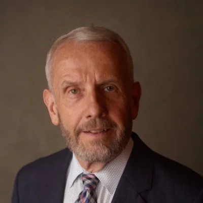 A portrait of an older man with gray hair and a beard, wearing a suit and tie, against a plain background.