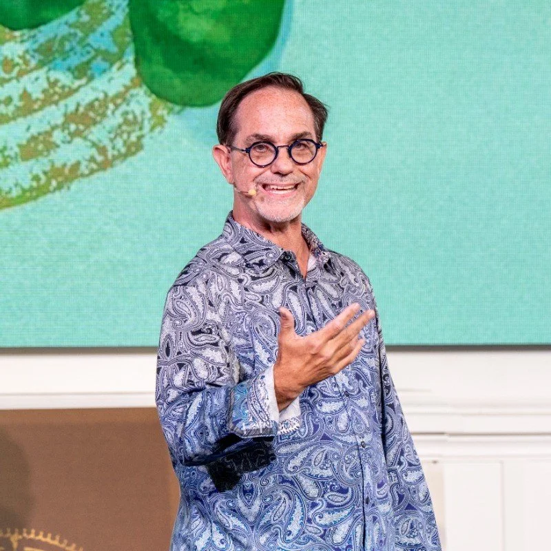 A man smiling, wearing glasses and a paisley-patterned shirt, speaking in front of a large screen with a green design.